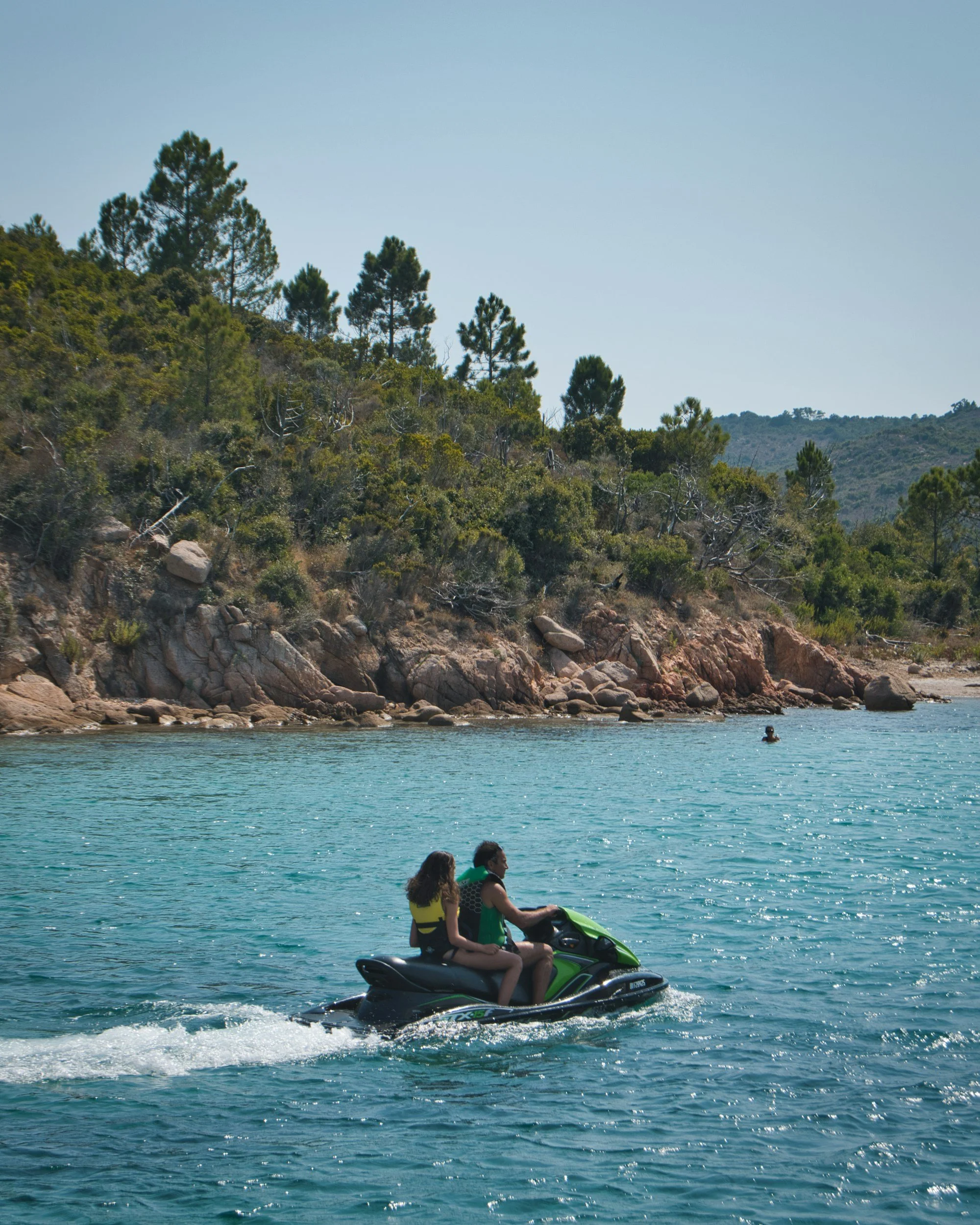Deux personnes en maillot de bain sur une deux-places jet ski dans un lac ou océan, avec une côte rocheuse et des arbres en arrière-plan, sous un ciel dégagé.