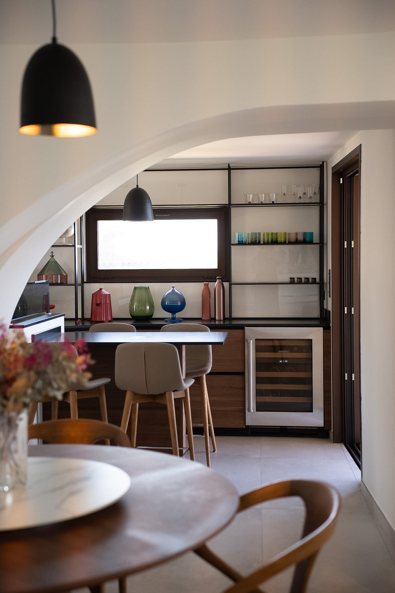 Intérieur d'une cuisine moderne avec un mur blanc, des étagères en métal contenant des verres et des tasses colorées, et un réfrigérateur intégré. Décorations colorées sur une table, ambiance chaleureuse et lumineuse.