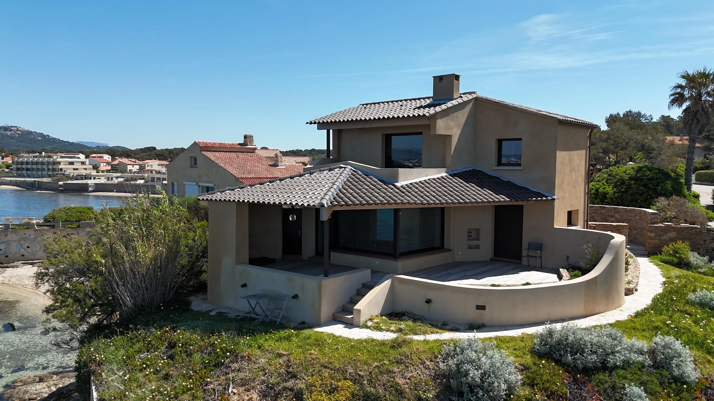 Maison moderne avec vue sur la mer, entourée de végétation, sous ciel bleu clair.