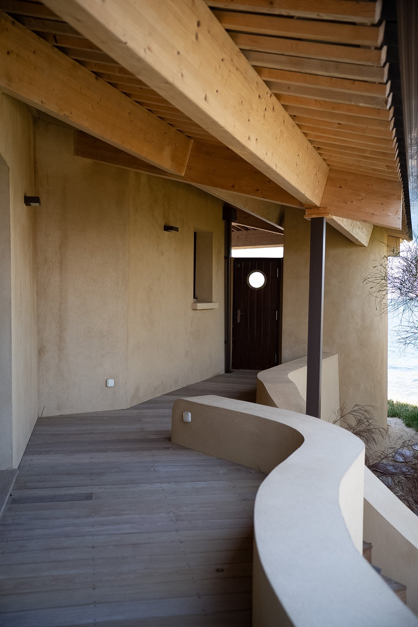 Une façade d'une maison moderniste avec un balcon en bois, un sol en bois, un mur beige, une porte en bois sombre avec un petit hublot, et un banc en béton avec une forme courbe.