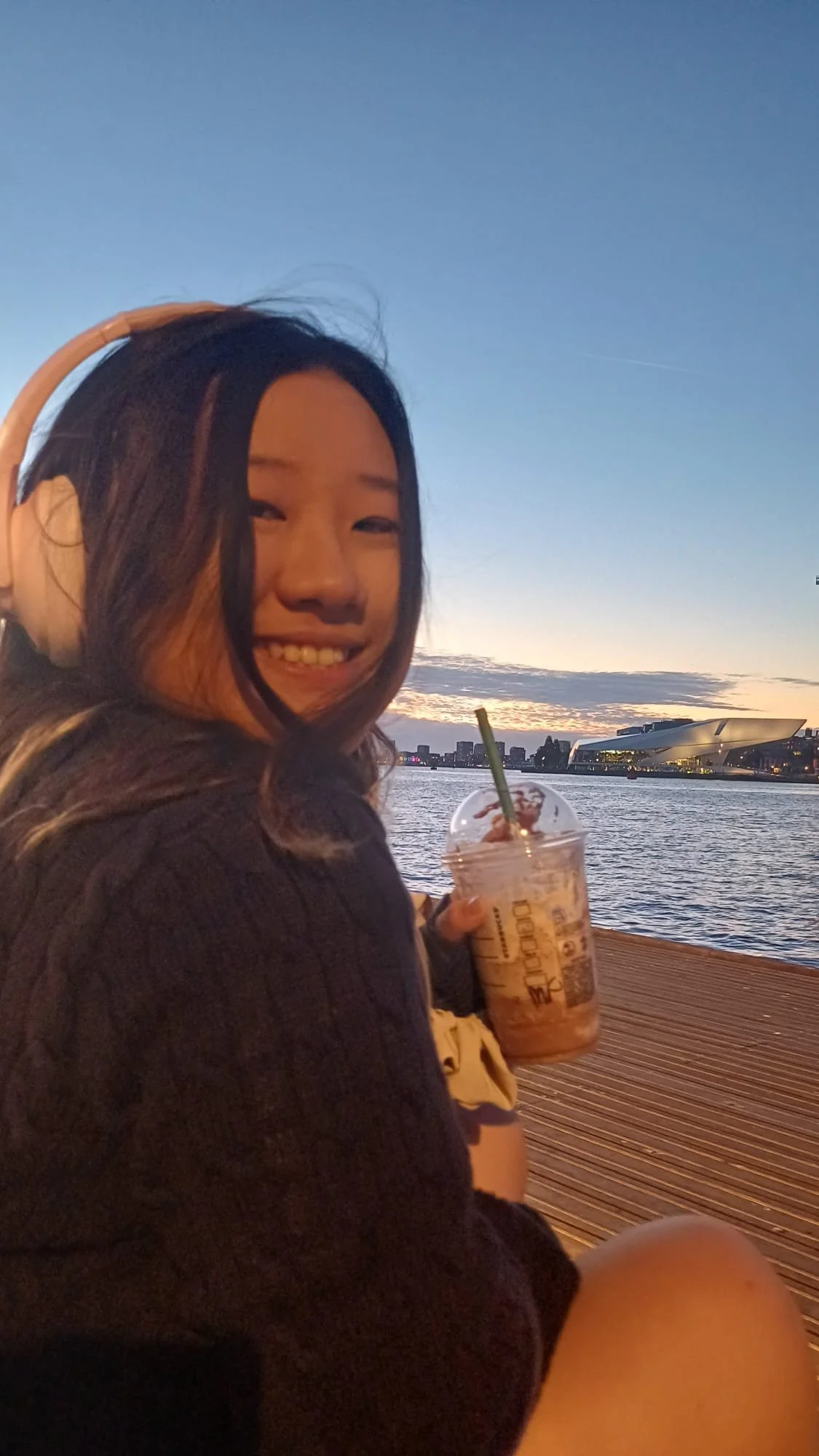 A young woman with long dark hair, wearing headphones and a black sweater, sitting by a river during sunset, holding a large iced coffee with whipped cream and a straw, smiling at the camera.