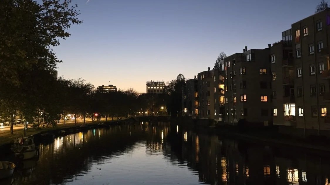 Dusk scene of a canal with calm water reflecting surrounding apartment buildings and trees, with some lights on in the apartments.