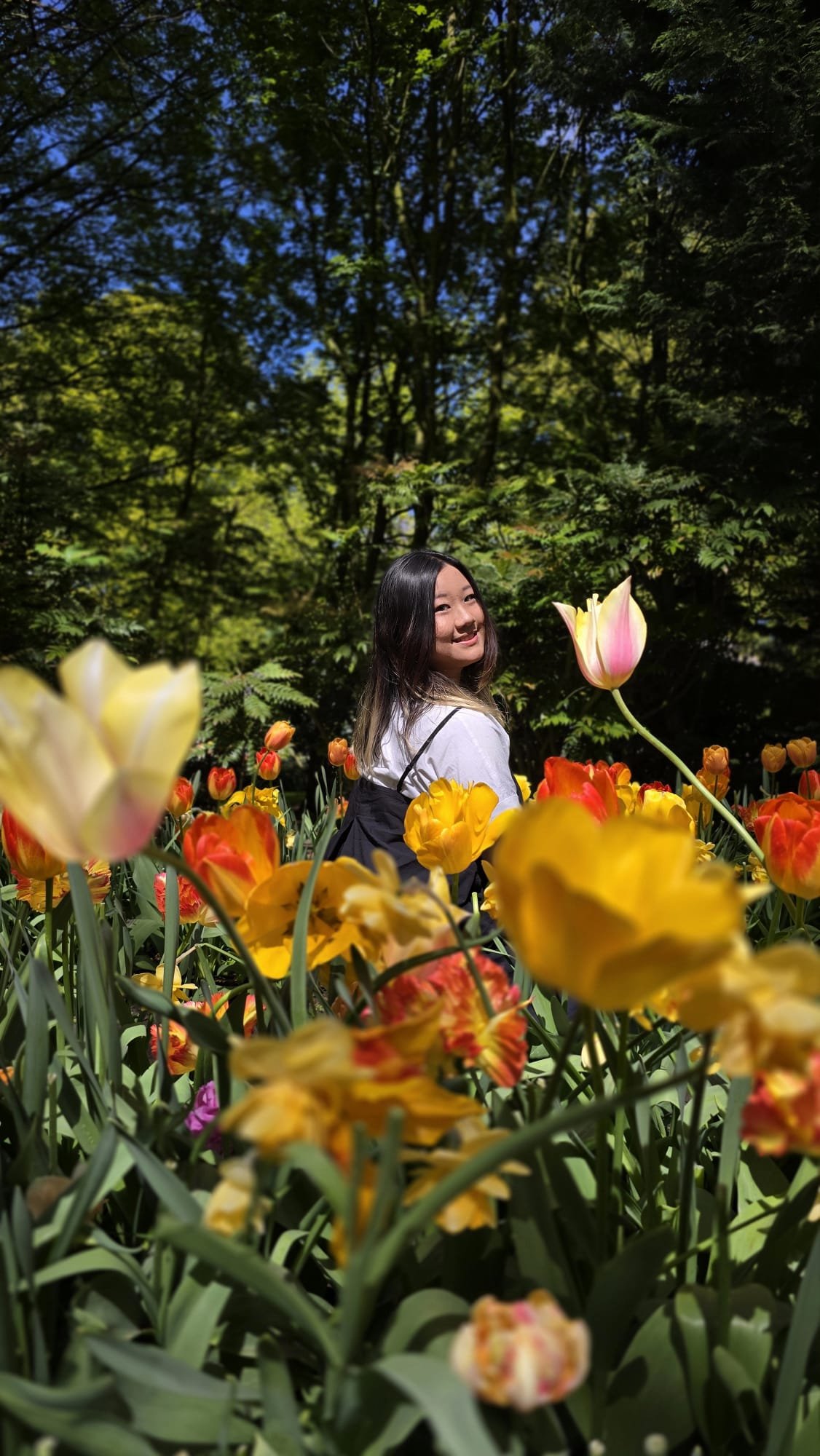 A young woman with long black hair smiling in a colorful flower garden with yellow, orange, and pink tulips, surrounded by green leafy trees under a blue sky.