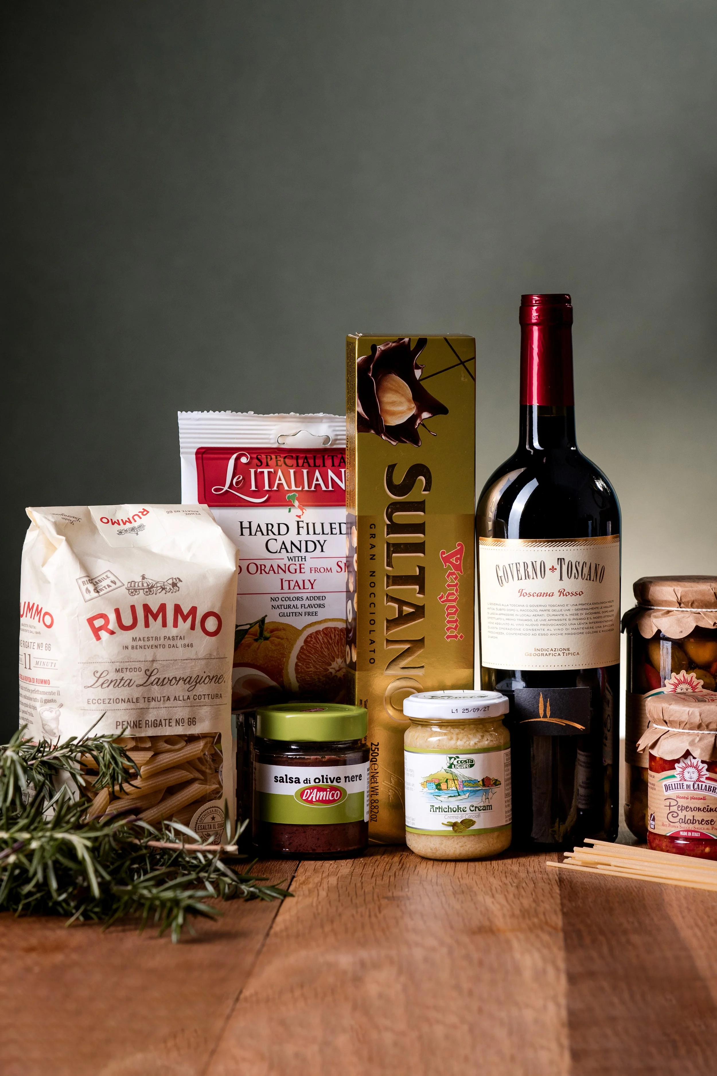 A variety of Italian food ingredients on a wooden surface, including dried pasta, a bottle of red wine, pasta sauce, artichoke cream, and packaged snacks, with some herbs on the side.