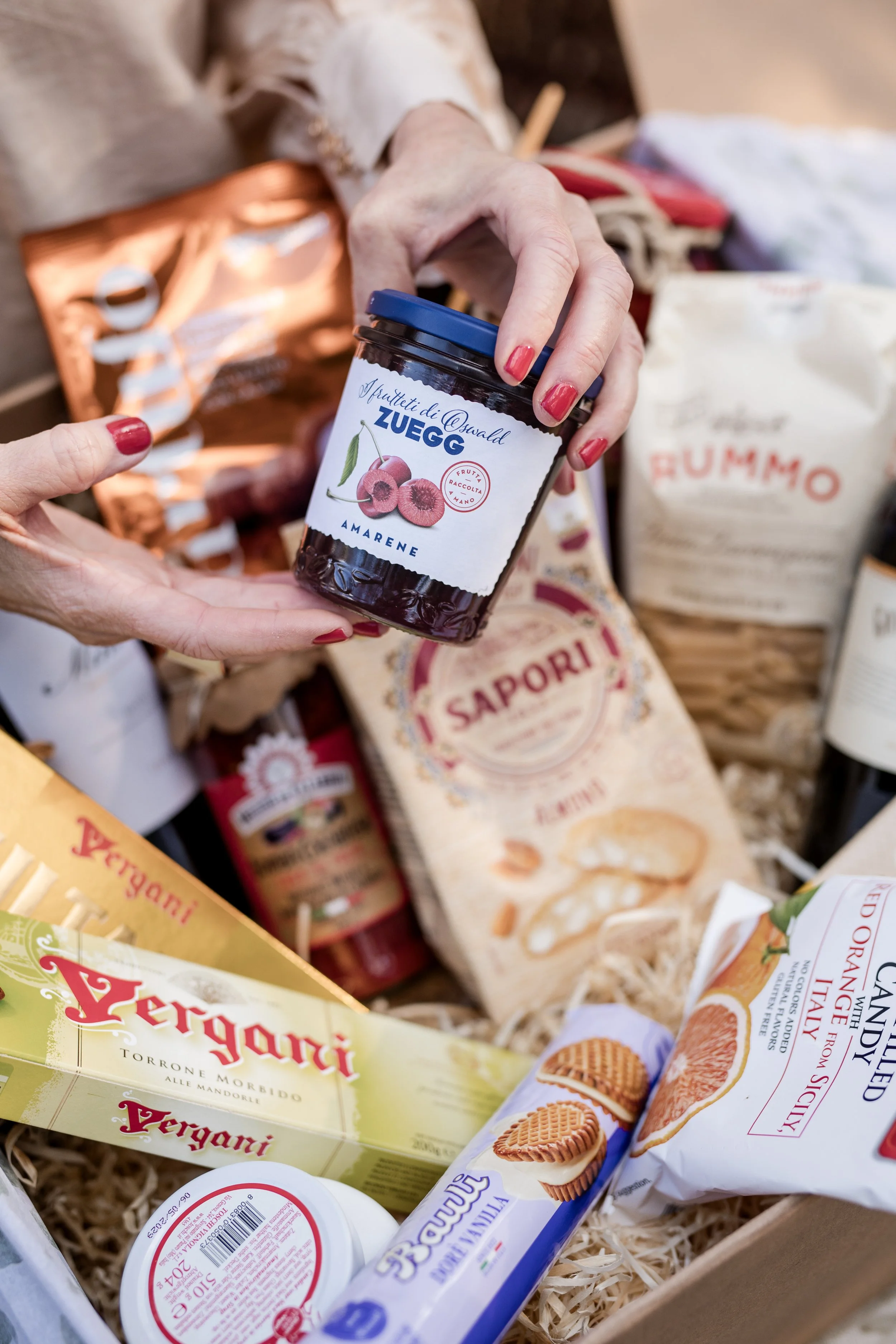 Close-up of a person's hand with red-painted nails holding a jar of Zuegg apricot fruit spread. In the background, various Italian food products are visible, including packages of Pirongia cookies and Rummo pasta, all on shredded paper.