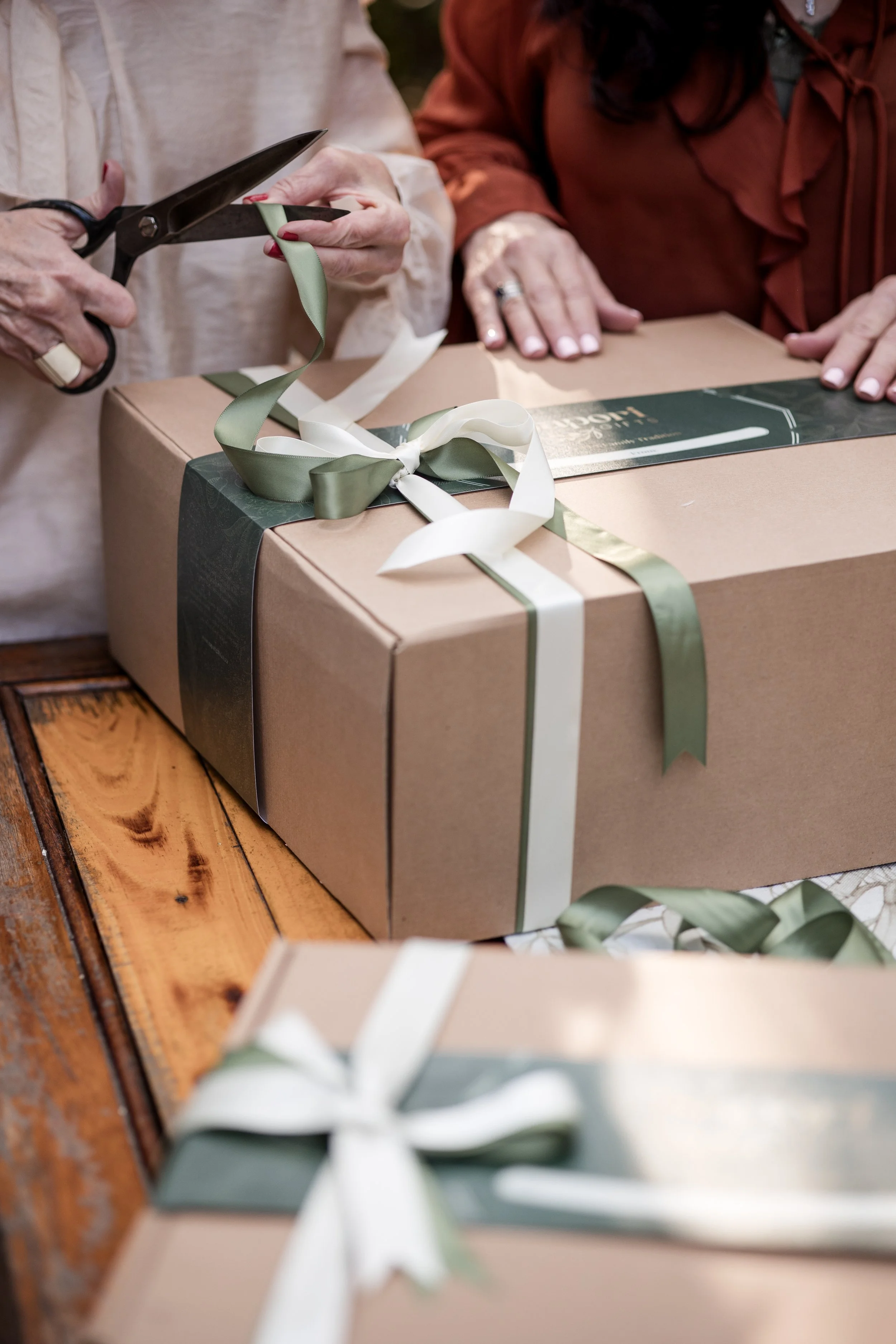 Close-up of two women wrapping a gift with brown paper, green and white ribbons, and scissors, on a wooden table.
