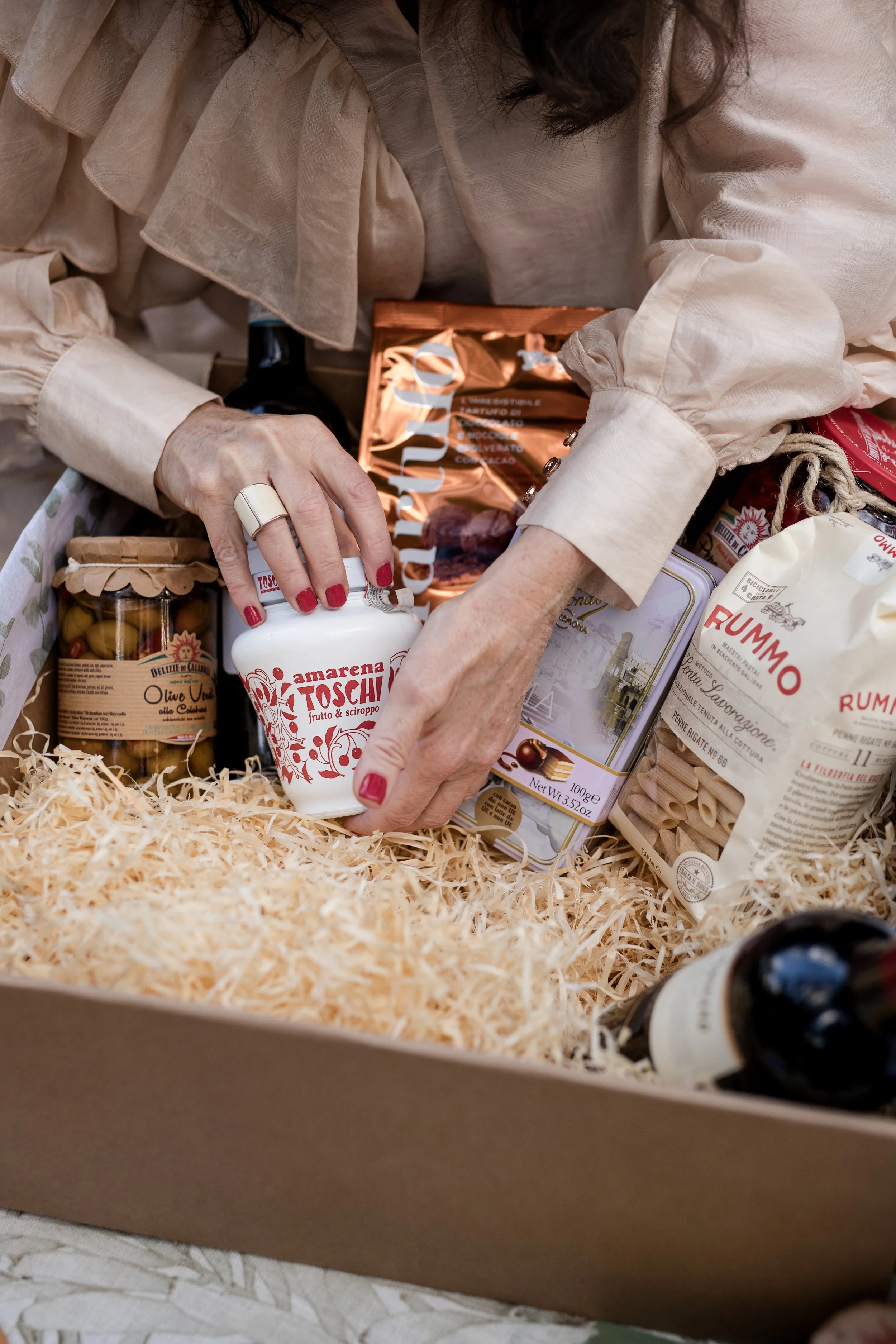 A person wearing a beige blouse with ruffled sleeves is reaching into a box of assorted food products. The box contains jars of preserved vegetables, packages of pasta, a bottle, and a bag of rice, all arranged on shredded paper filler.