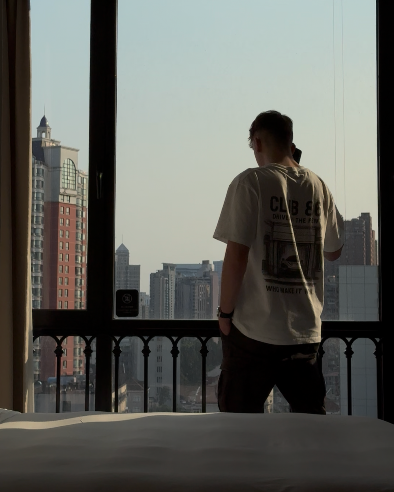 A person standing on a balcony, looking out at a city skyline during the daytime, with high-rise buildings visible in the background.