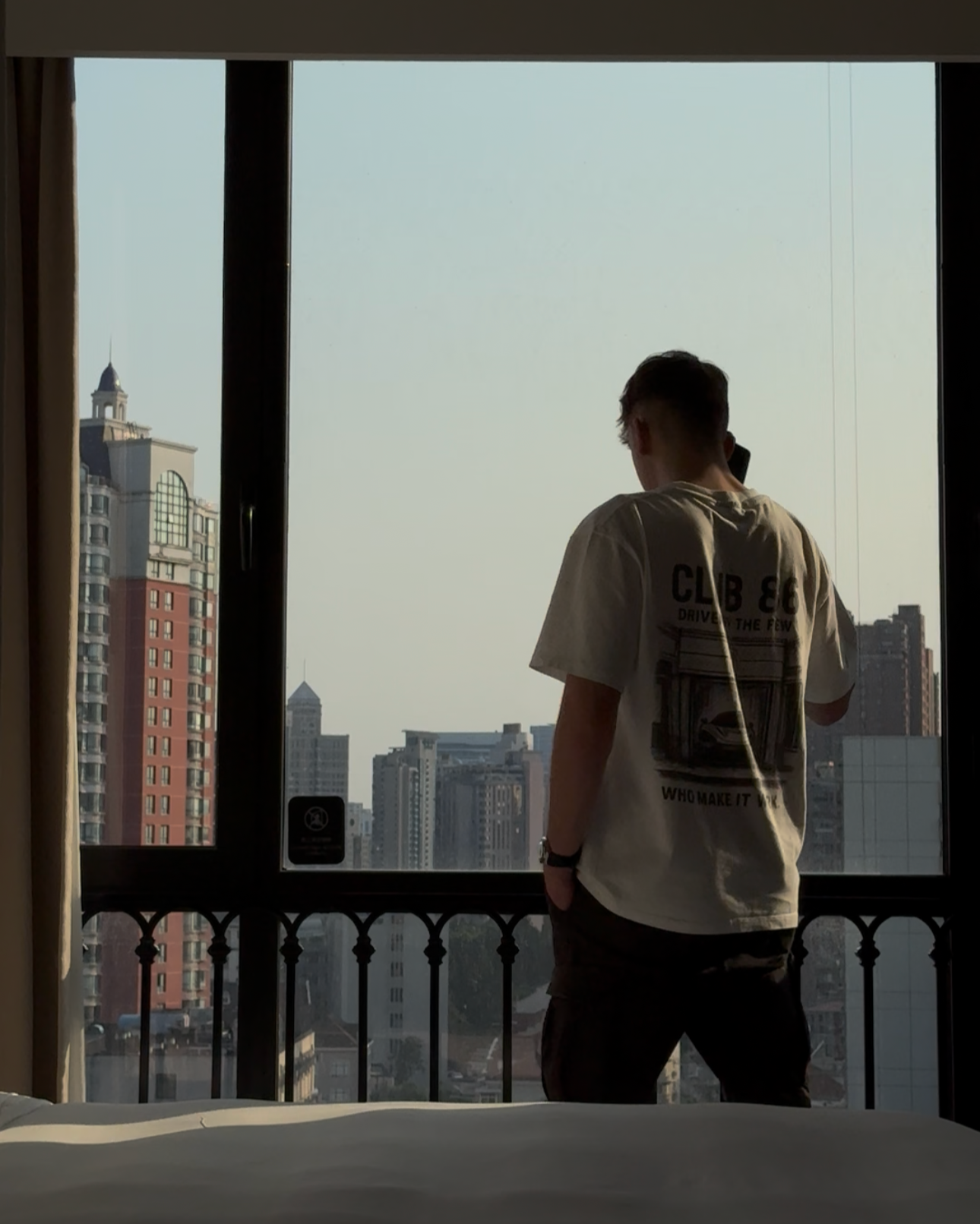 A person standing on a balcony overlooking a city skyline with tall buildings during sunset, facing away from the camera.