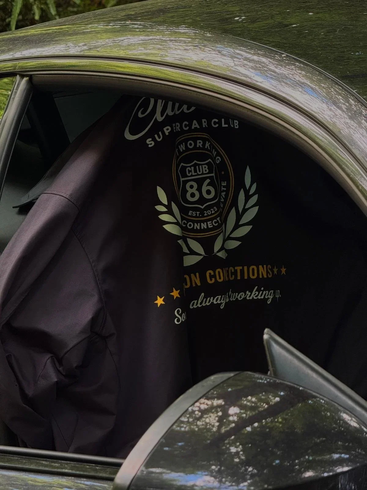 Car window showcasing a large black banner with white and gold text and design, including a badge with '86' and surrounding leaves, partially covered by the car's window frame and reflects green trees and sky.