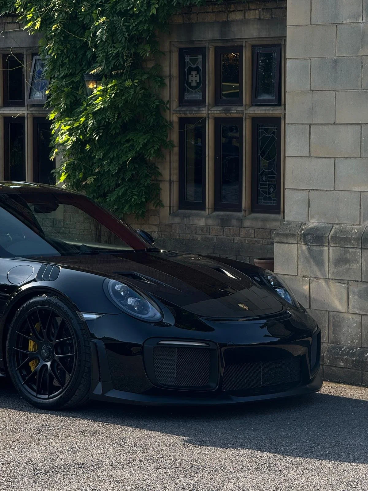Close-up of a black sports car parked in front of a stone building with a window and green shrubbery.