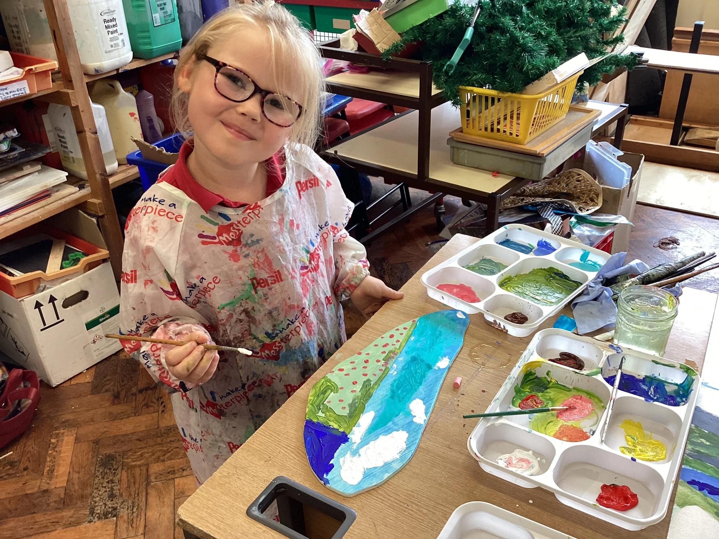 A young girl with glasses and a paint-splattered apron smiling while holding a paintbrush. She is in an art studio or classroom with painting supplies, an unpainted Christmas tree in a yellow basket, and various art materials around her.