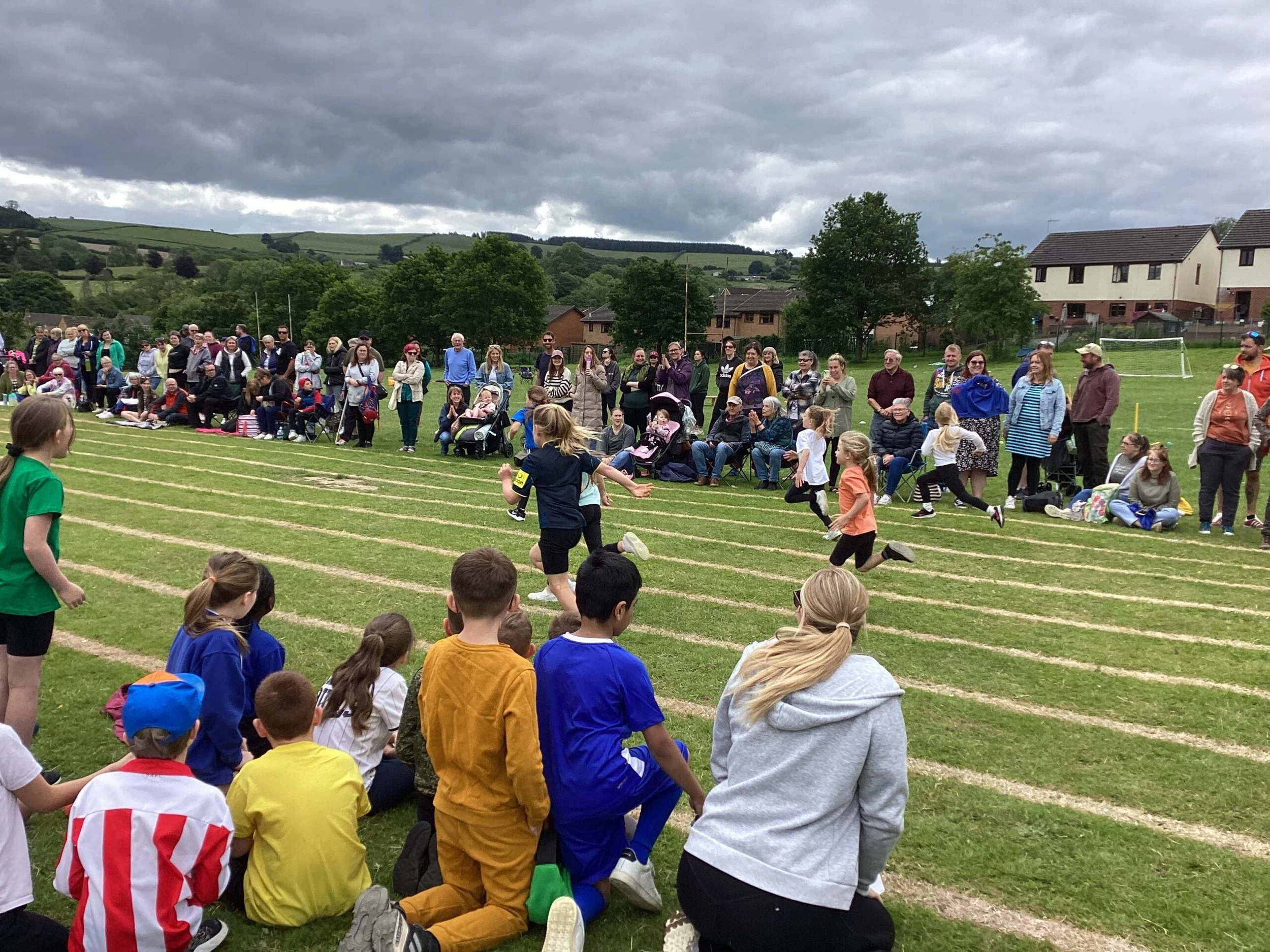 Group of children and adults gathered on a grassy sports field, some sitting and some standing, with trees, houses, and a cloudy sky in the background, participating in an outdoor event or sports day.