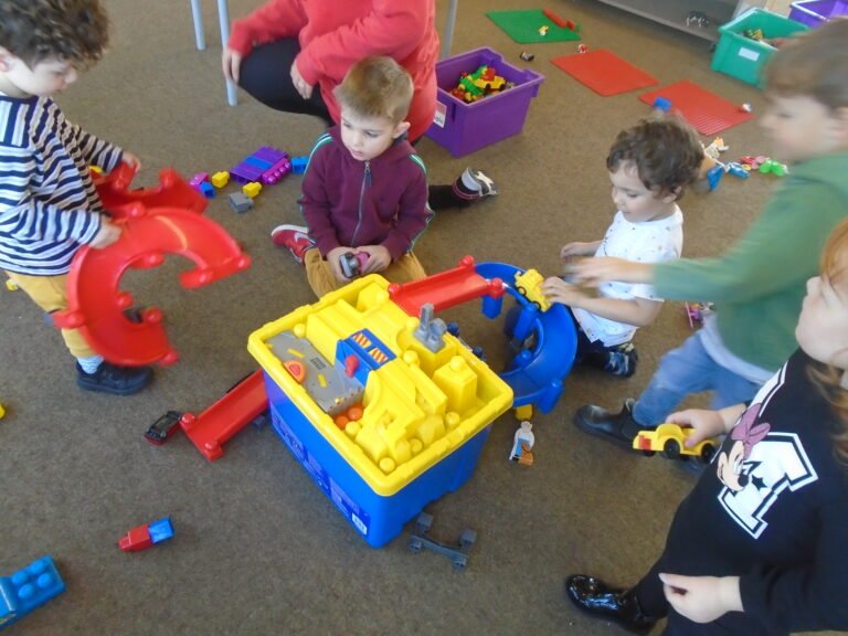 Children playing with a large blue and yellow toy construction set on the carpeted floor, surrounded by various smaller toys and building pieces.
