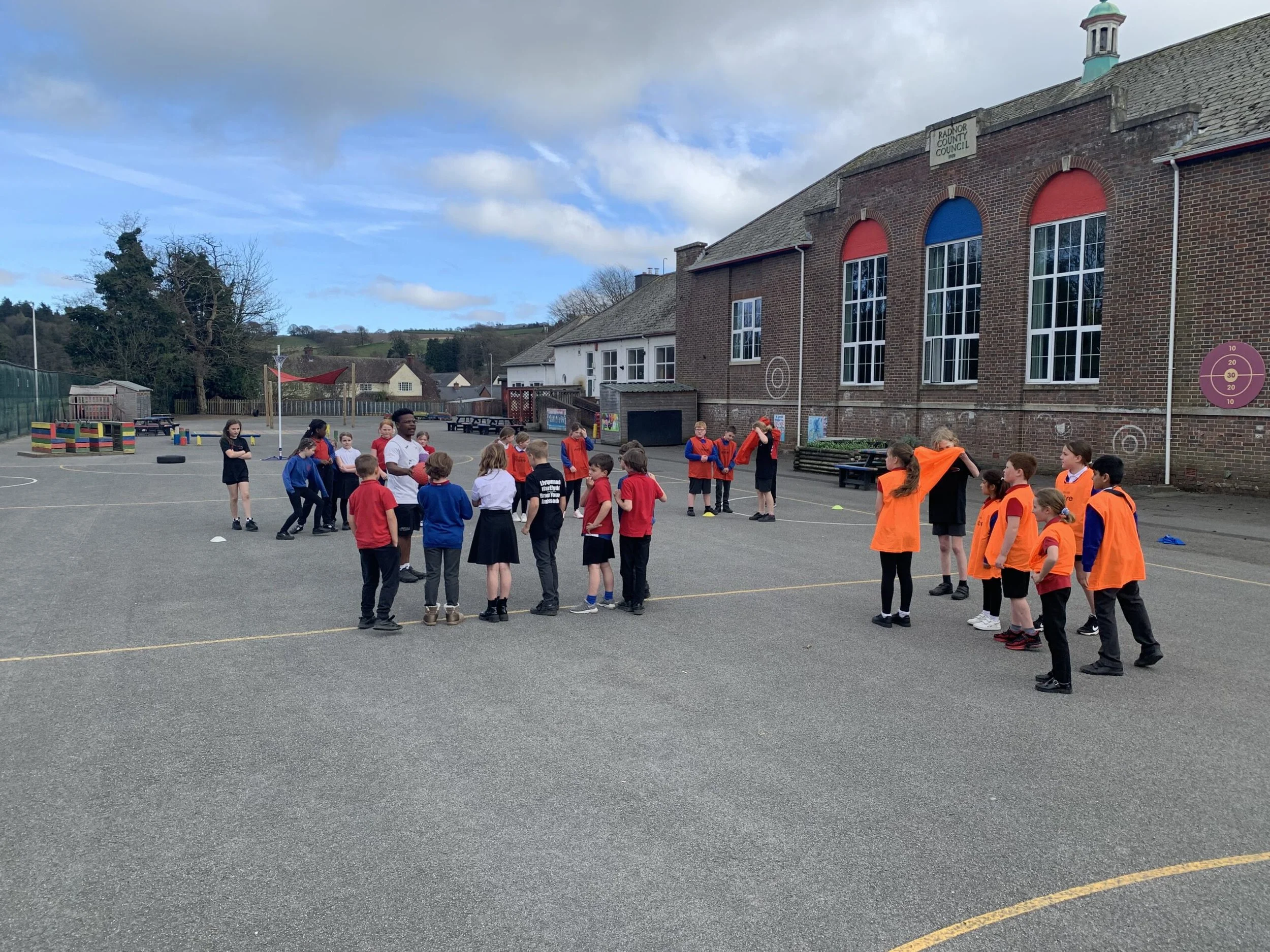 Children and a teacher gathered outside a school building with large windows, some wearing orange safety vests, during daytime. The school building has a brick facade and a sign reading 'Radnor County Council'. There are trees, hills, and a cloudy sky in the background.