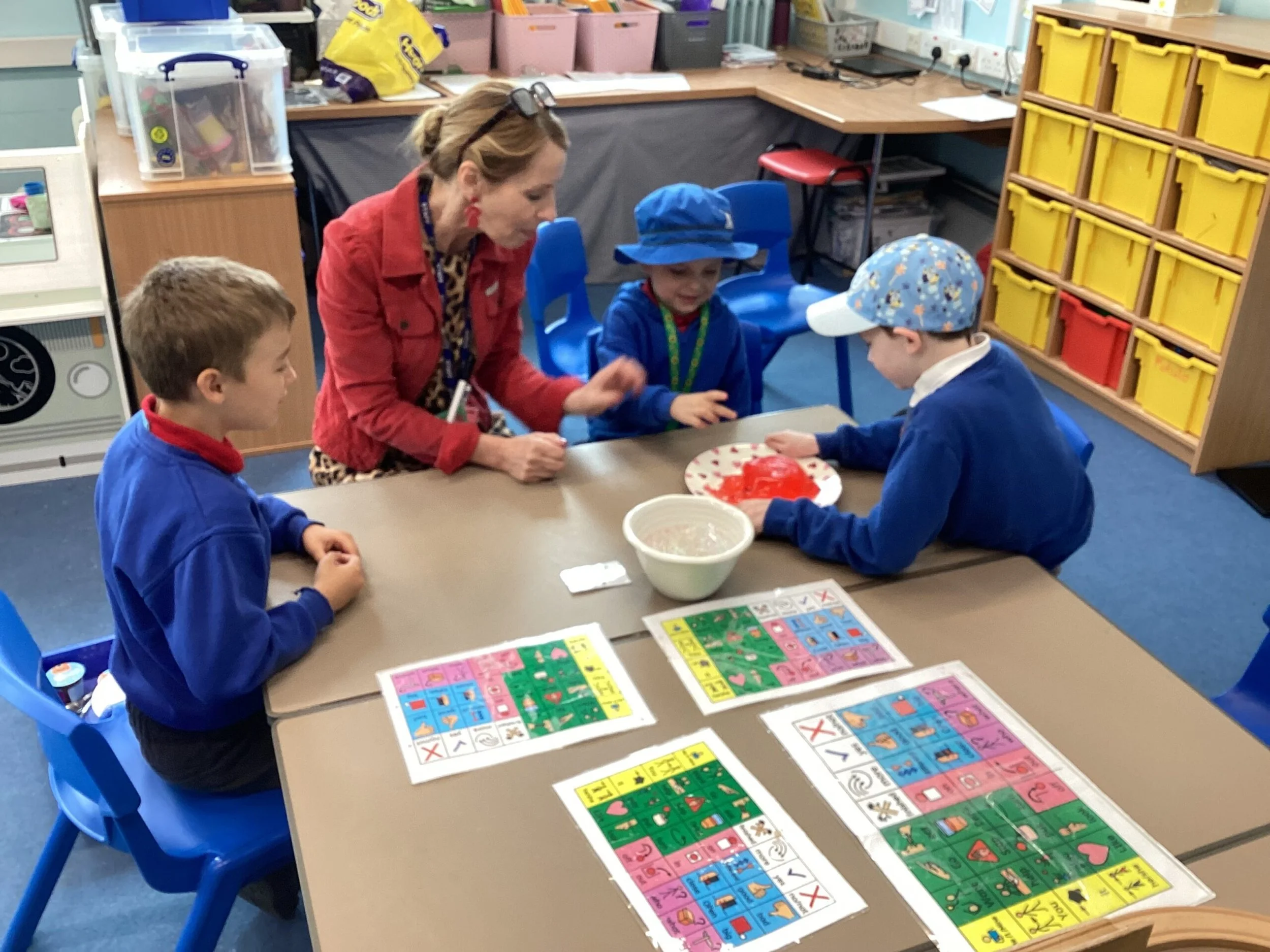 A teacher with glasses on her head and earrings, wearing a red jacket, is sitting at a table with three children in a classroom. The children are wearing blue uniforms and hats, and are engaged in a learning activity involving colorful alphabet and picture cards spread on the table. The classroom contains storage bins, educational materials, and a shelf with yellow and red bins.