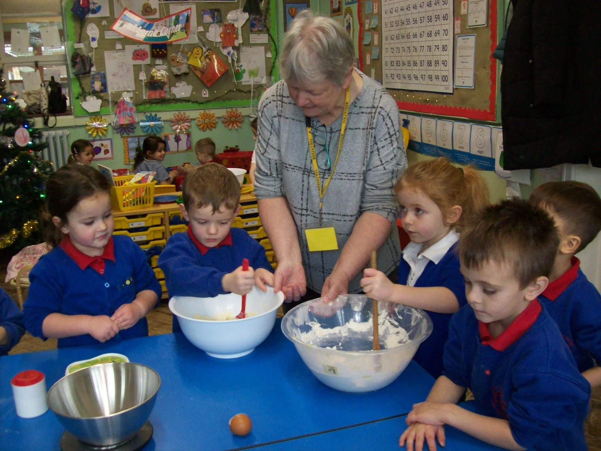 A group of young children wearing blue and white uniforms gathered around a table in a classroom, helping an elderly woman mix ingredients in a large white bowl with a red spatula. The classroom has colorful decorations, charts, and artwork on the wa