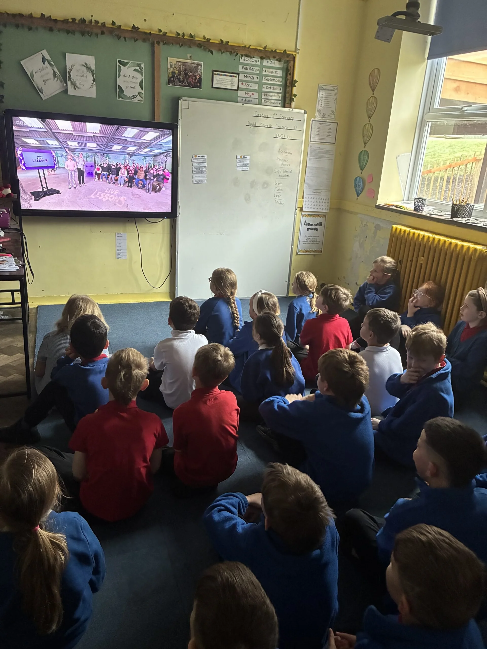 A classroom with children sitting on the floor watching a television screen. The television displays a scene labeled "Live Lessons." There is a whiteboard and various educational posters and materials on the walls, and sunlight is coming through a window.
