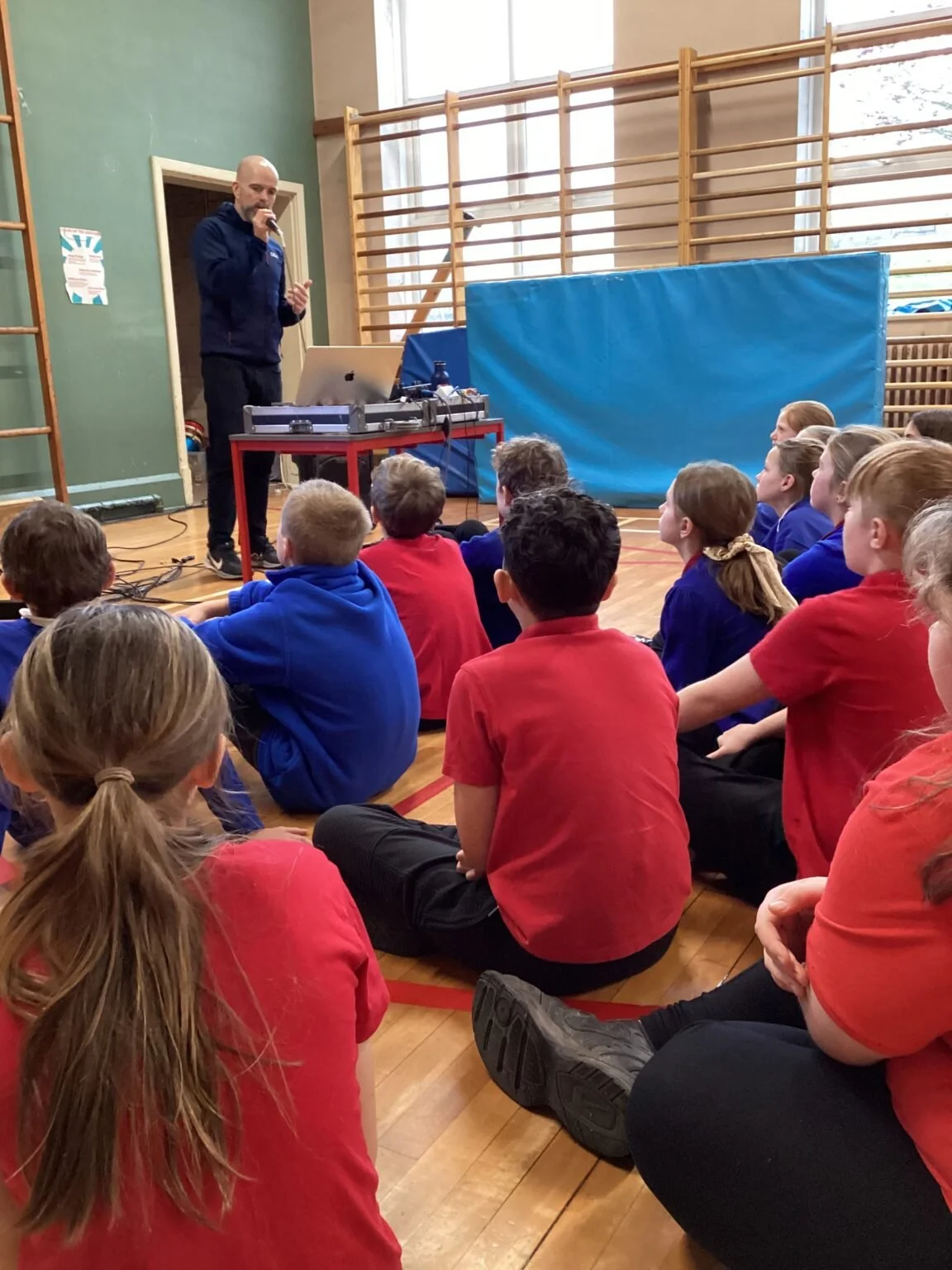 A man giving a presentation to a group of students sitting on the gymnasium floor. The man is standing at a table with a microphone, laptop, and a DJ controller. The students are in colorful clothing, mostly red and blue, and are attentively listenin