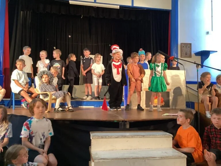 Children performing a play on stage during a school event, with some dressed in costumes like a Christmas hat, green dress, and casual attire, in a school auditorium.