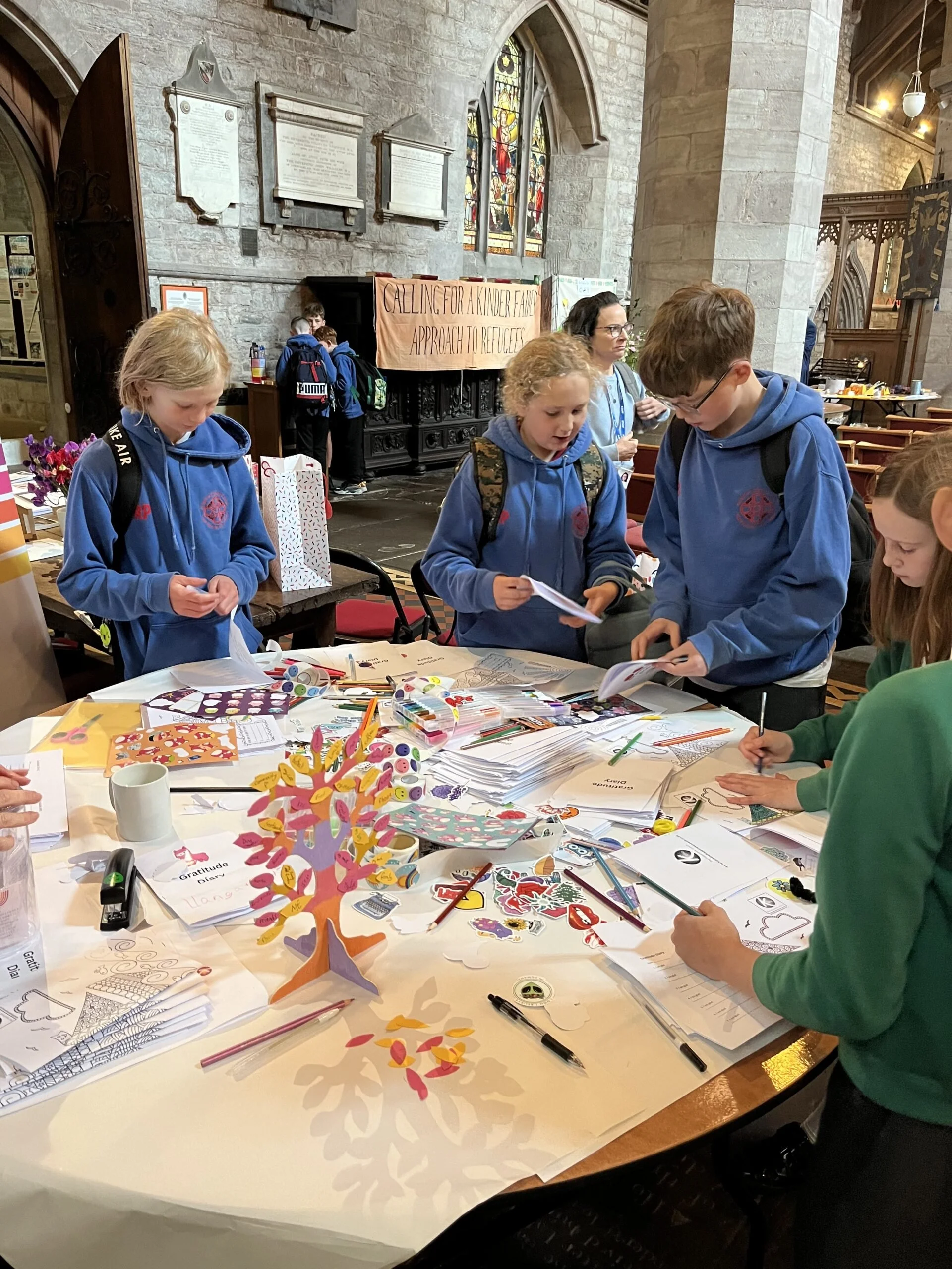 Children in blue hoodies gathered around a table inside a church, engaging in arts and crafts activities such as coloring and cutting paper. The table is filled with craft supplies, paper cutouts, and decorations. The church's interior features stain