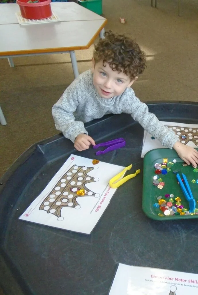 A young boy with curly hair working on a crown-shaped craft project at a table. There are small colored jewels and tools around him.