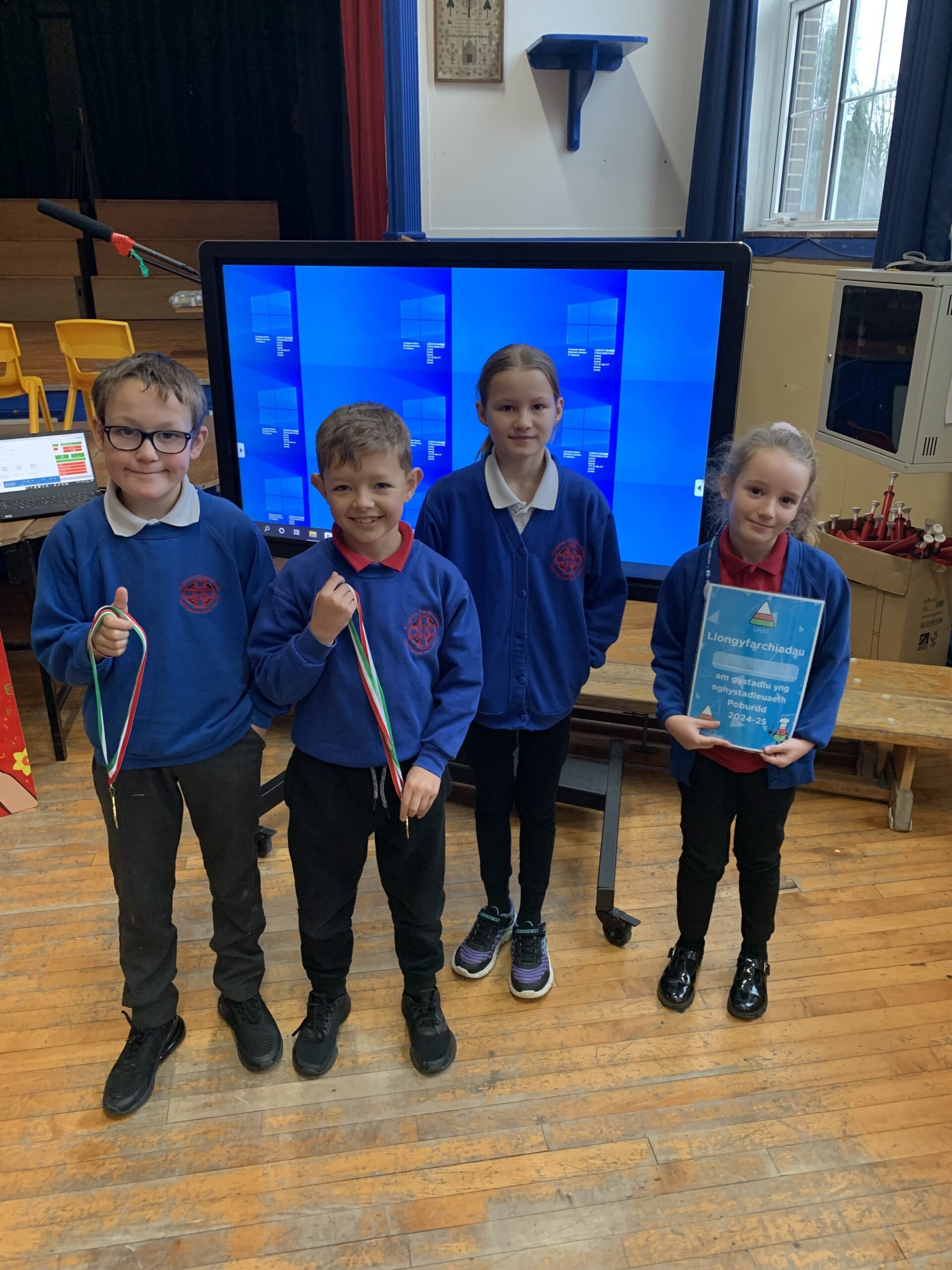 Four children, two boys and two girls, standing indoors in front of a computer screen, holding medals, smiling, wearing blue uniforms.