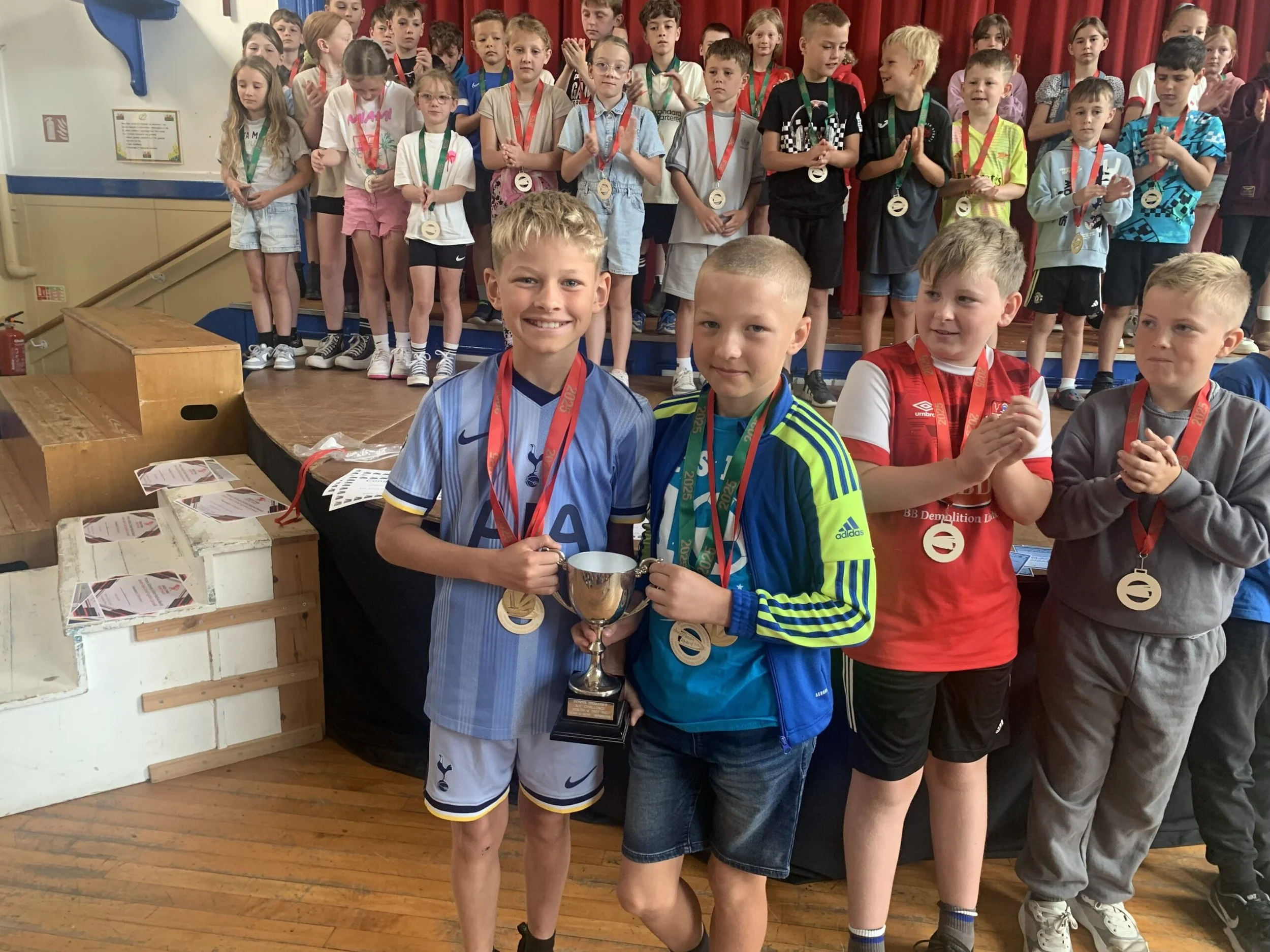 Children at an award ceremony, with two boys in the front holding a trophy, and the rest of the children standing behind, some wearing medals around their necks, on a stage with red curtains.