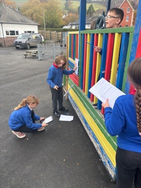 Four children in blue school uniforms practicing dialogues with scripts on a colorful playground, one standing inside the play structure, one writing on paper on the ground, and two reading from scripts.