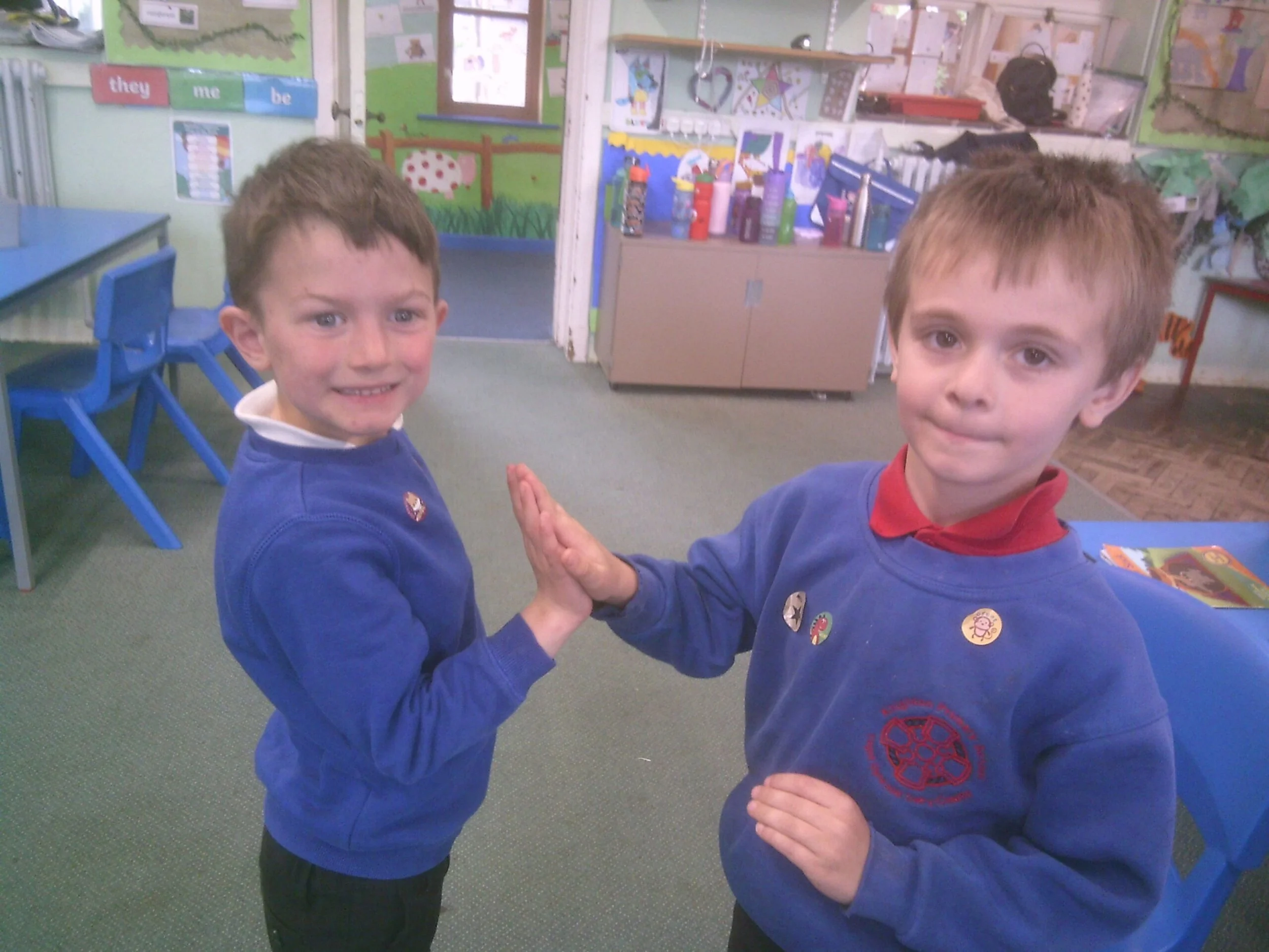 Two young boys in a classroom giving each other a high five. They are wearing blue sweaters, one with a red collared shirt underneath and the other with a white shirt. The classroom background includes colorful decorations, books, and tables.