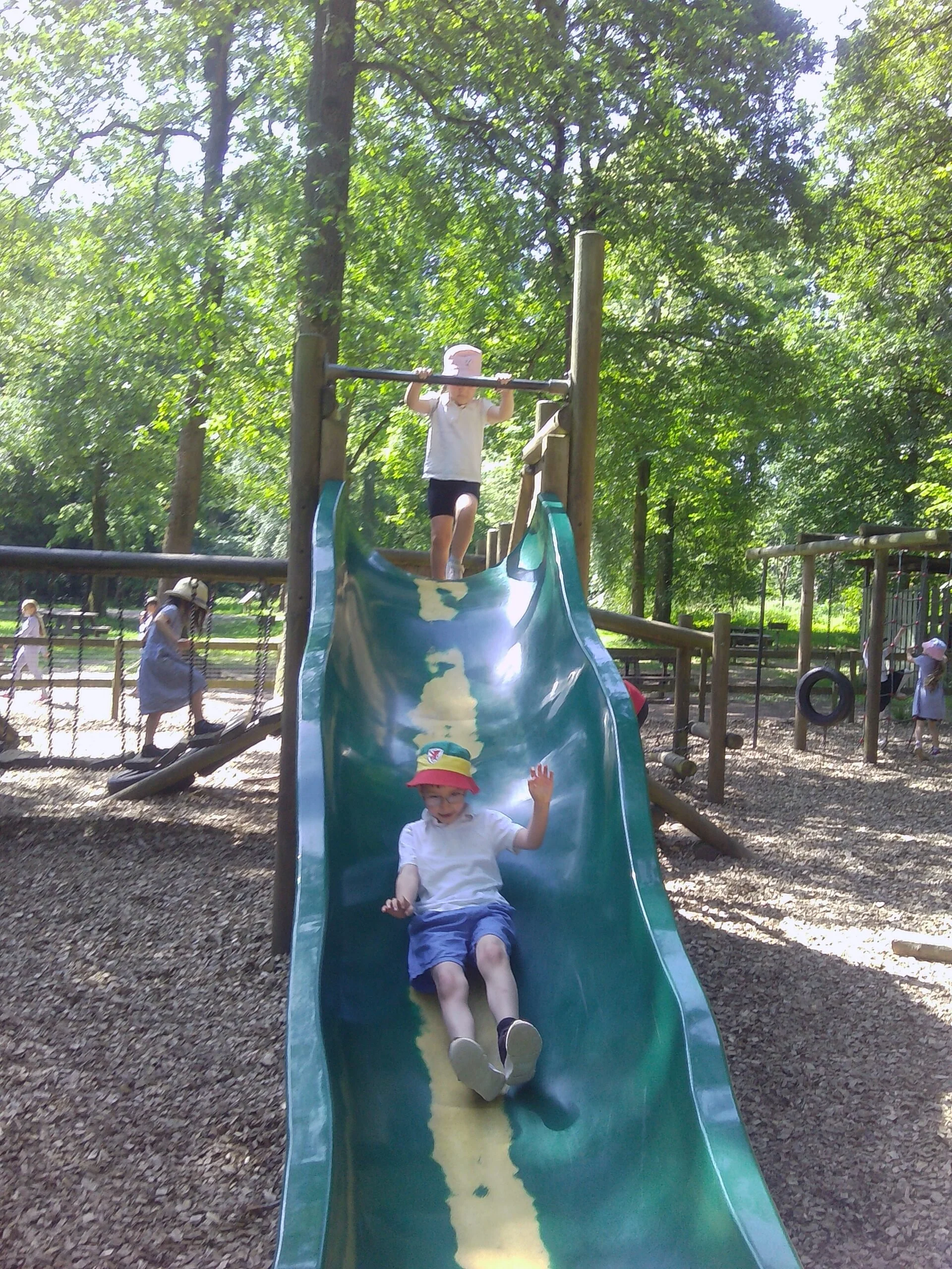 Children playing on a slide in a park surrounded by trees. Two children are sliding down, one at the top holding onto the bar and the other at the bottom waving.