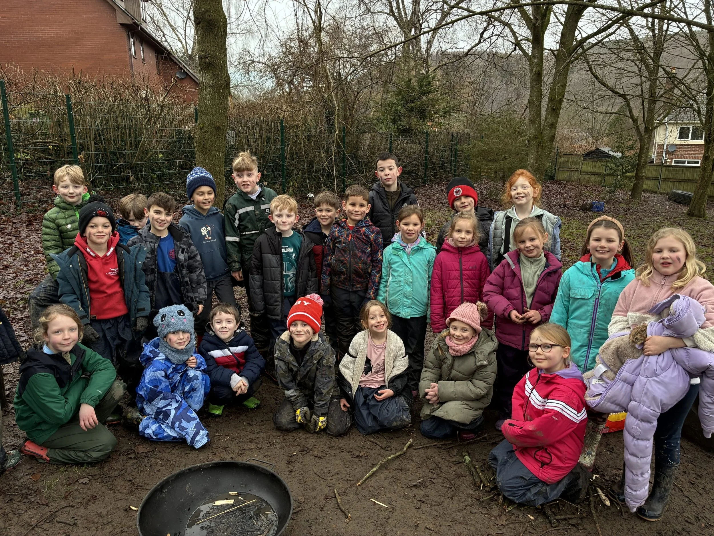 A group of children, wearing colorful outdoor jackets and hats, posing outdoors in a muddy area with trees and residential houses in the background.