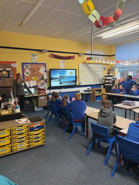 Classroom with students seated at blue desks, teachers at the front, and a large interactive screen displaying the ocean and a yellow submarine.