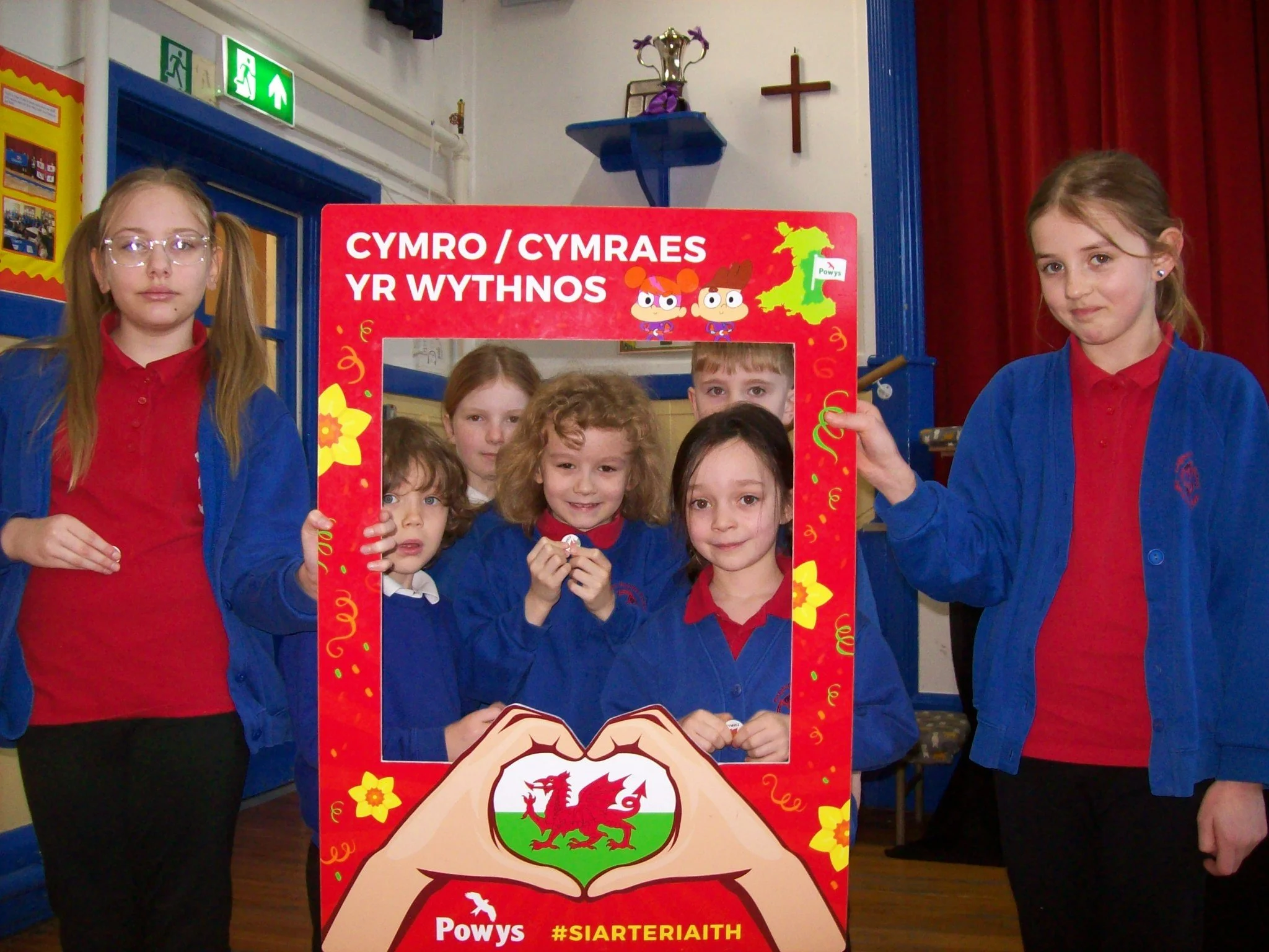 Group of children and two adults posing behind a red frame with Welsh language text and symbols, inside a hall with a cross and religious items on the wall.