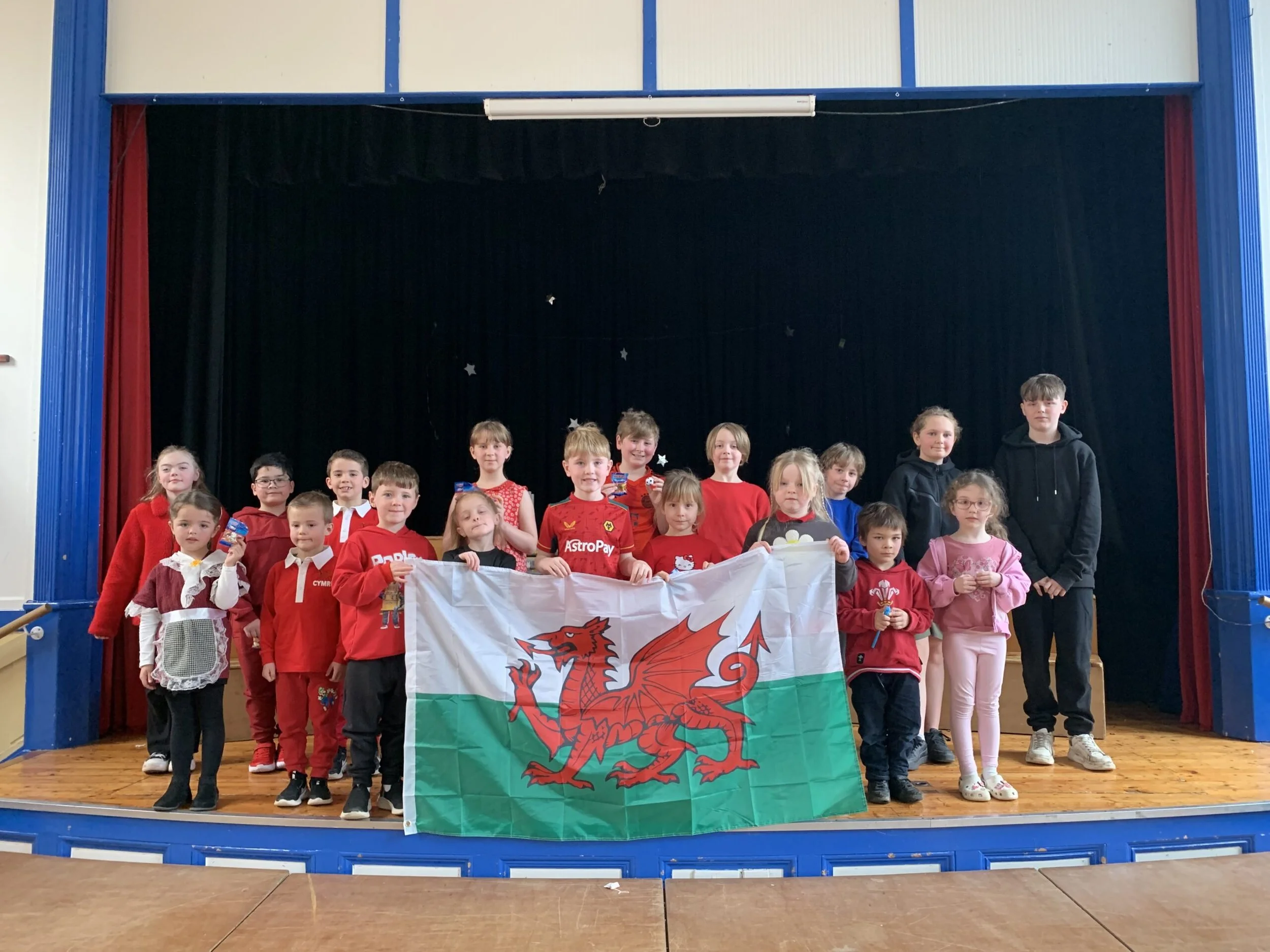 A group of children on a stage holding a large Welsh flag with a red dragon, in front of a black curtain with star decorations, inside a blue and red auditorium, with some children wearing red clothing.