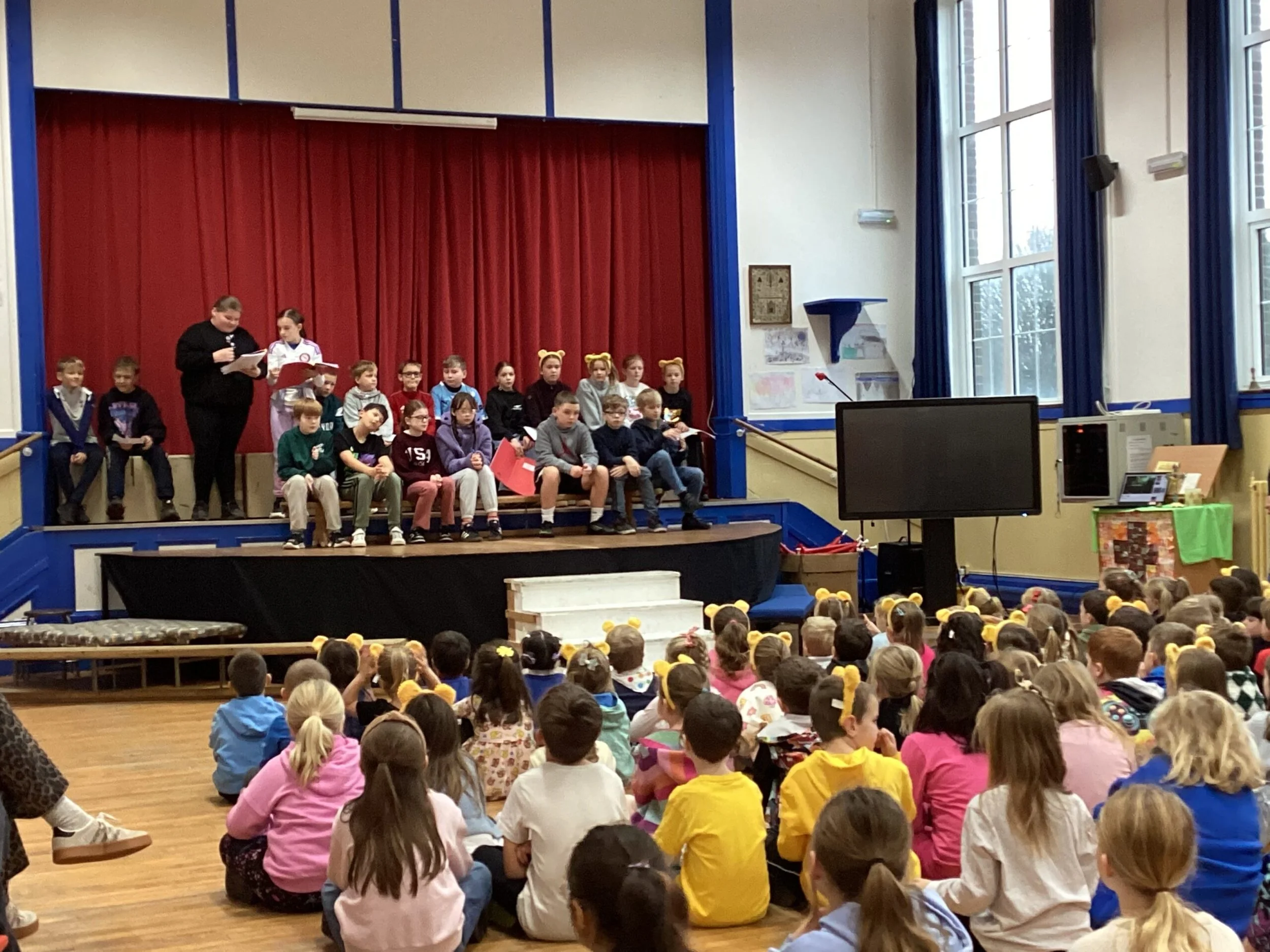 Children gathering in a school auditorium for a presentation or performance on stage with red curtains, and a woman speaking to the audience. Many children are seated on the floor, facing the stage, with some wearing yellow headbands. The room has la