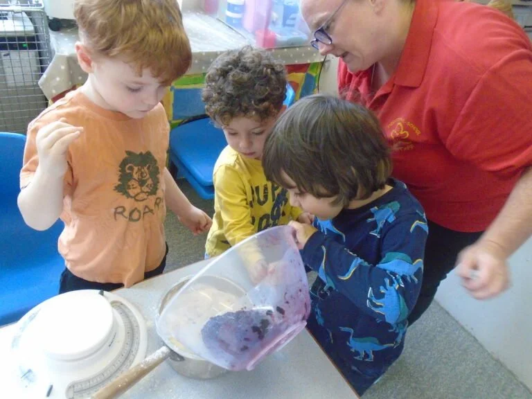 An adult woman and three young children gathered around a table, looking into a clear container with purple contents, possibly slime or a craft project, in a classroom setting.