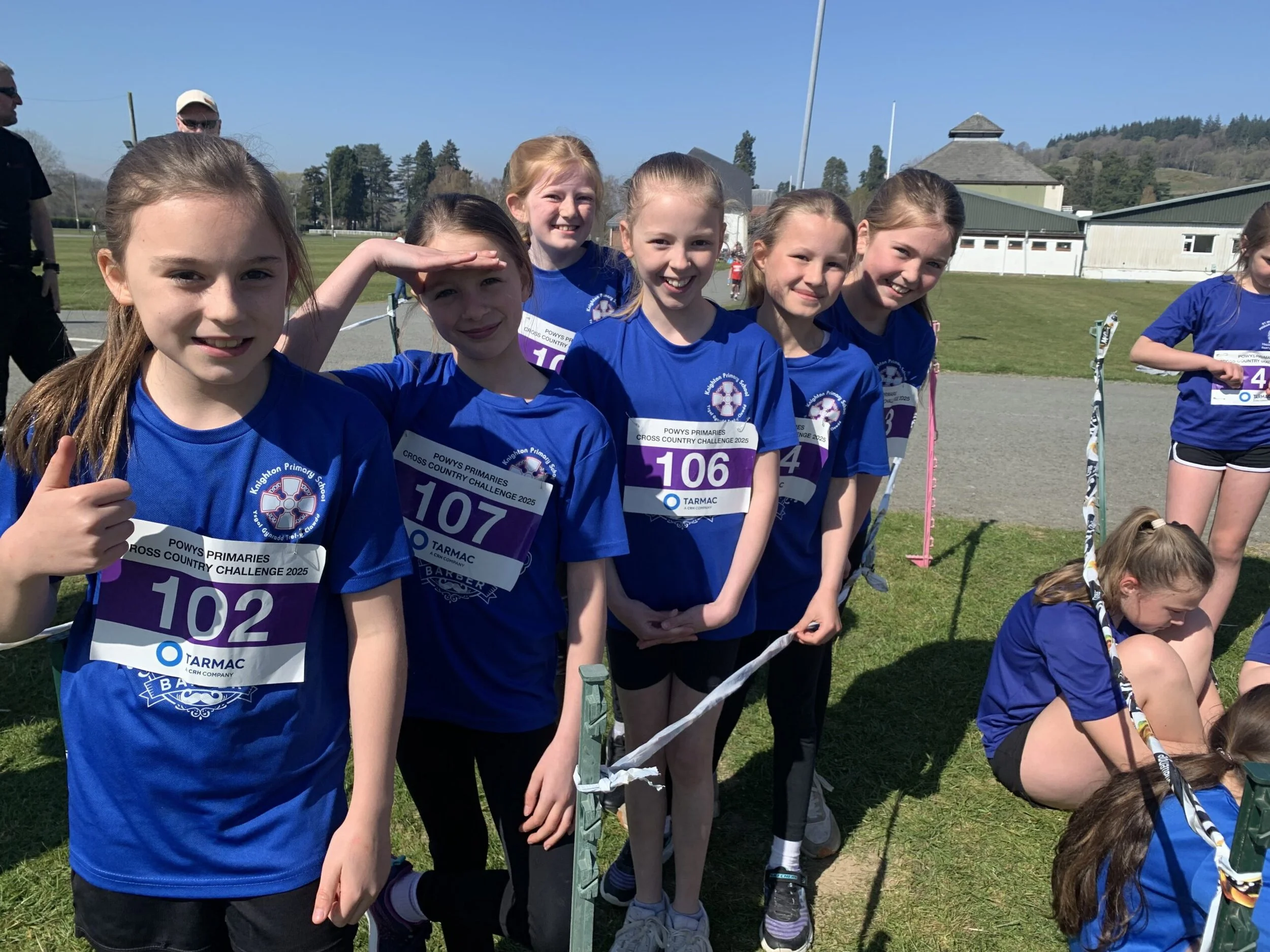A group of young girls in blue sports uniforms standing in a line outdoors, smiling, with some holding race bibs, participating in a cross country event on a sunny day.