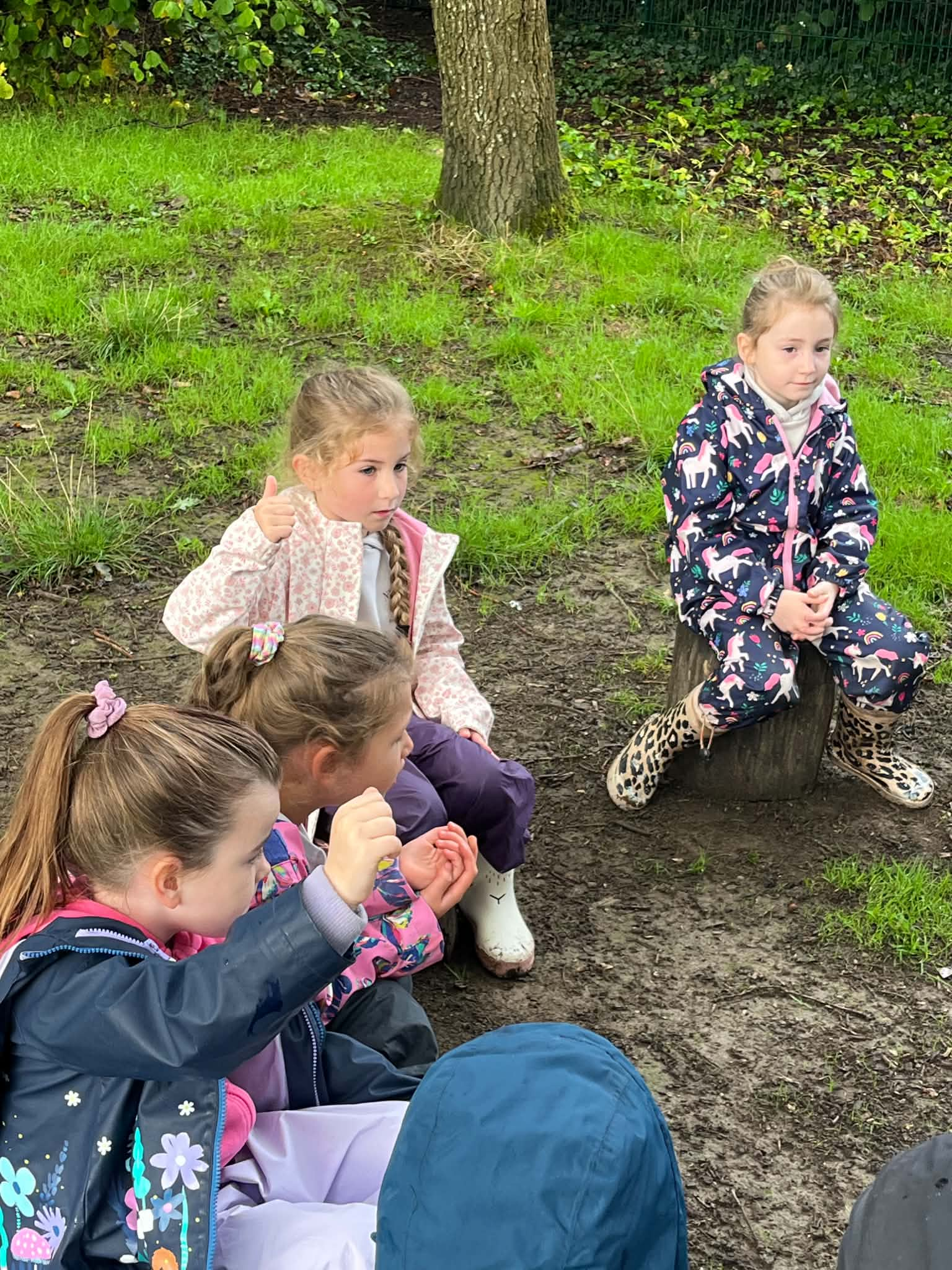 A group of children sitting outdoors on the ground near a tree, with one girl sitting on a small animal figure with painted hooves, and others sitting nearby, some with their hands near their faces.