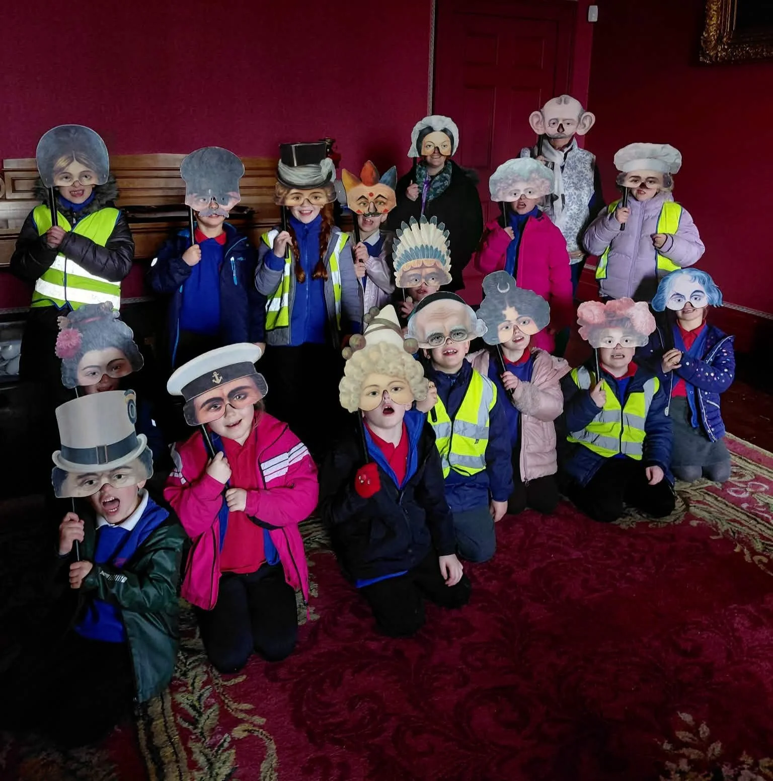 A group of children with some adults behind them, all wearing masks of different characters and holding sticks with masks attached, posing indoors against a red wall with ornate gold picture frames.