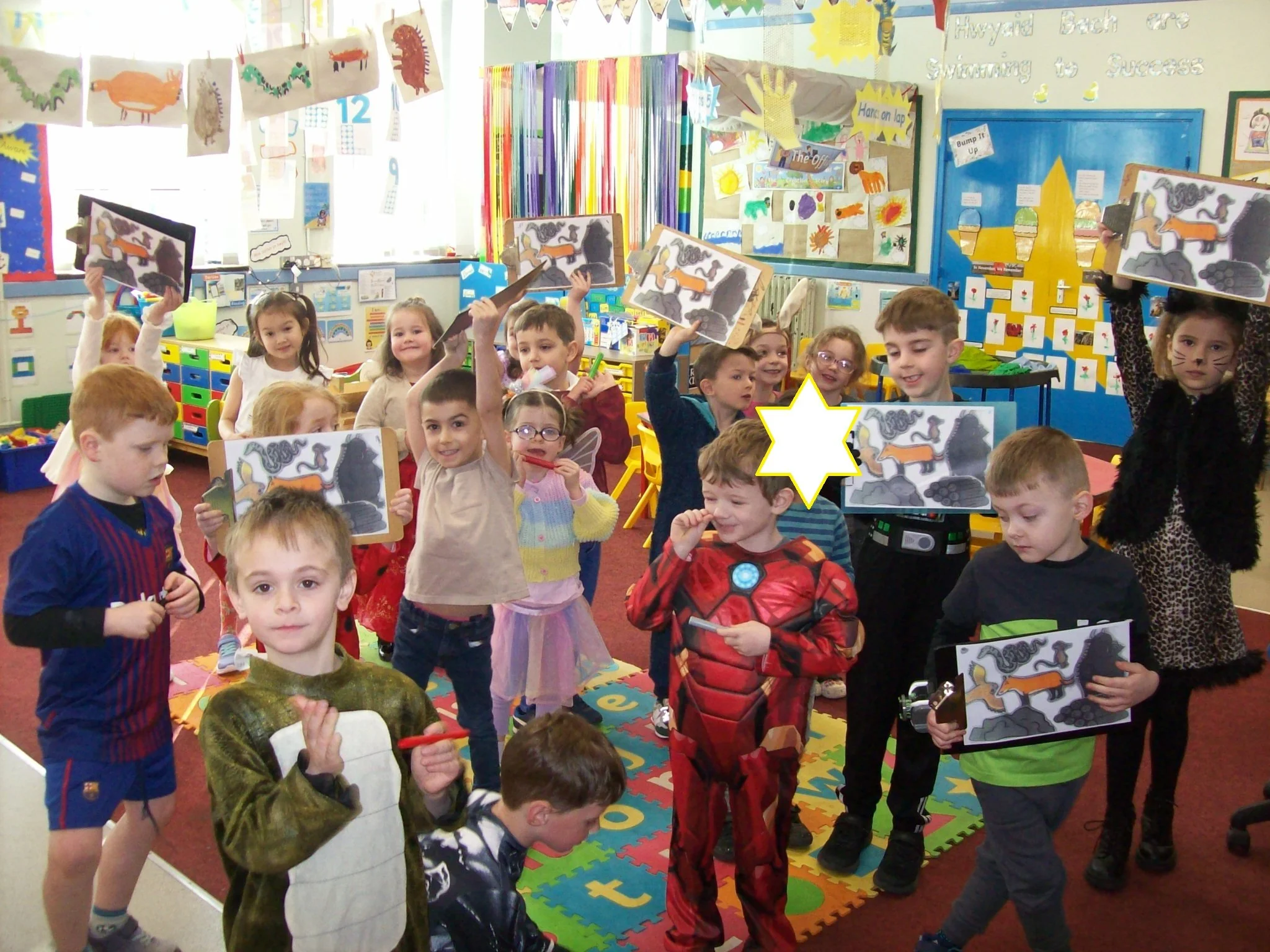 A classroom filled with young children, some dressed in costumes like a fairy, Spider-Man, and animal pajamas, holding up drawings of dinosaurs, with colorful decorations on the walls.