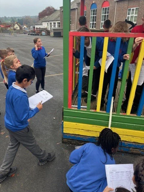 Group of school children outside, some holding clipboards and stationery, gathered around a colorful play structure with vertical bars, with a school building in the background.