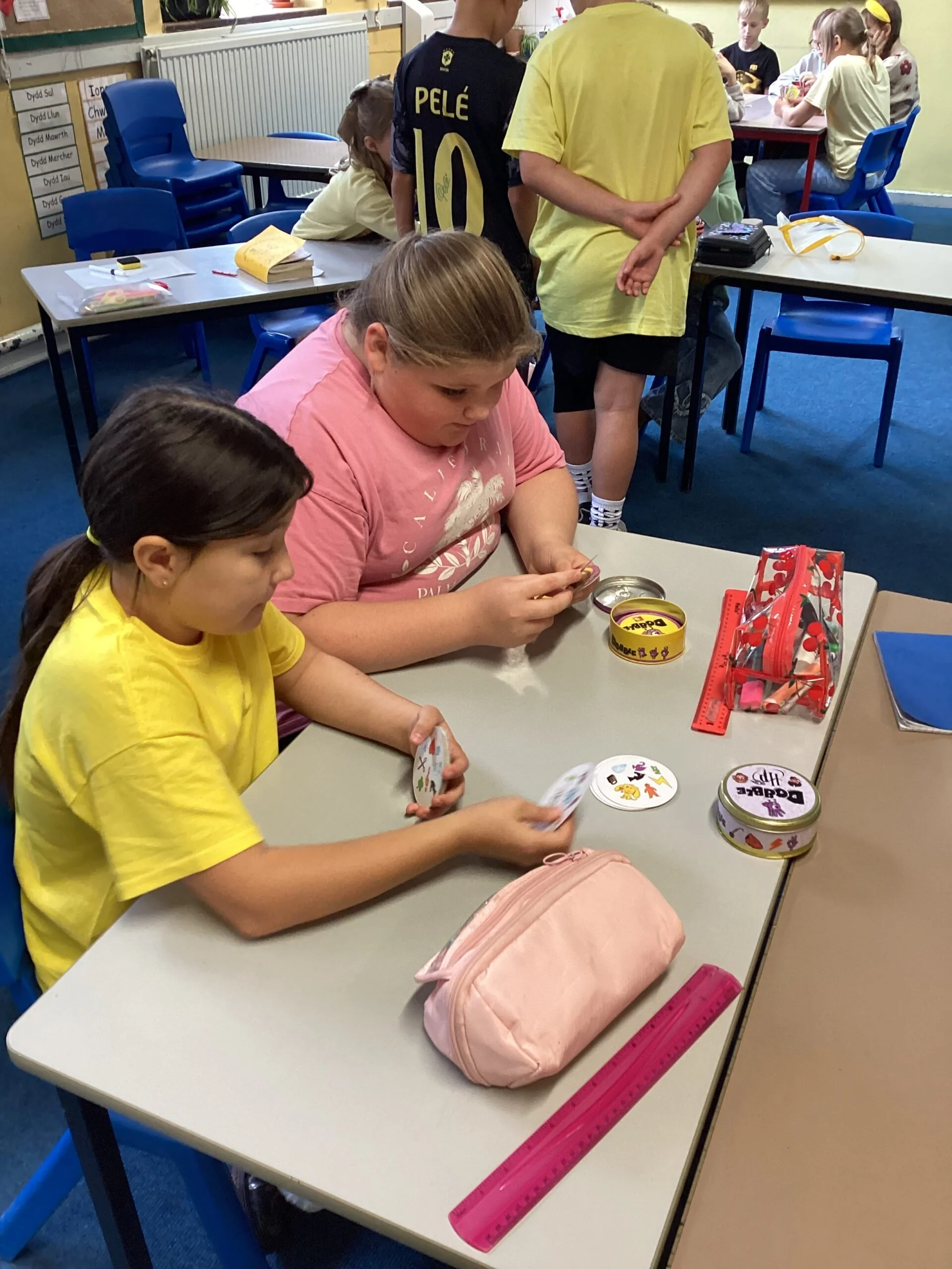 Children in a classroom sitting at desks, some are playing cards, others are working or talking, with school supplies and colorful objects on the desks.