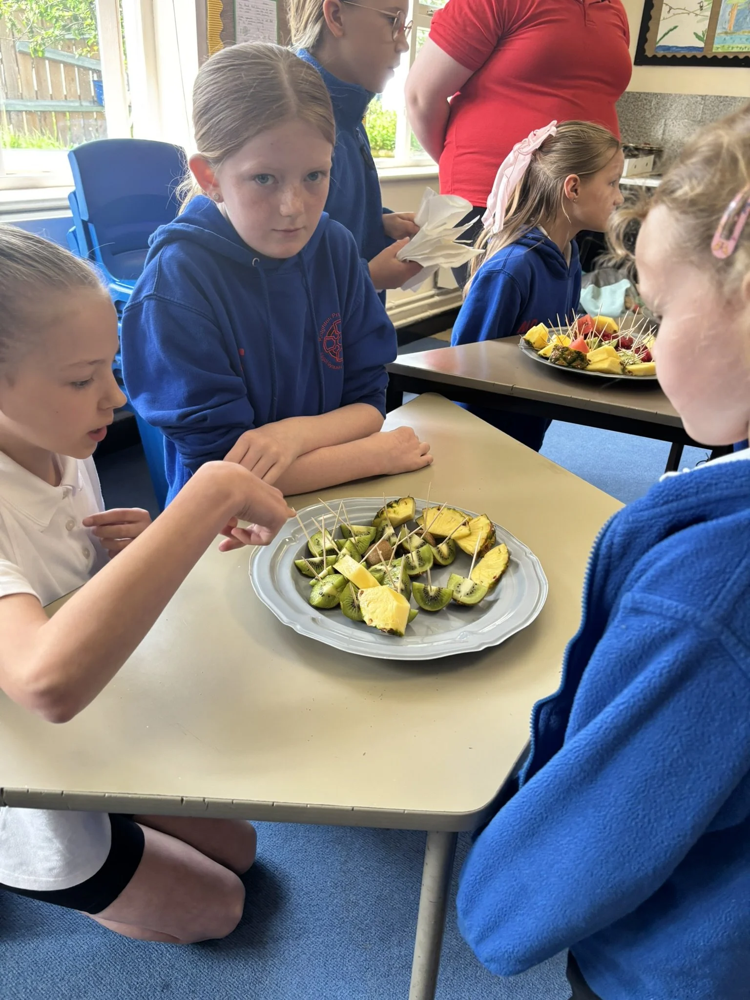 A group of children, some with freckles, are sitting at a table in a classroom, looking at trays of sliced kiwi, pineapple, and strawberries with toothpicks. An adult is standing behind them.