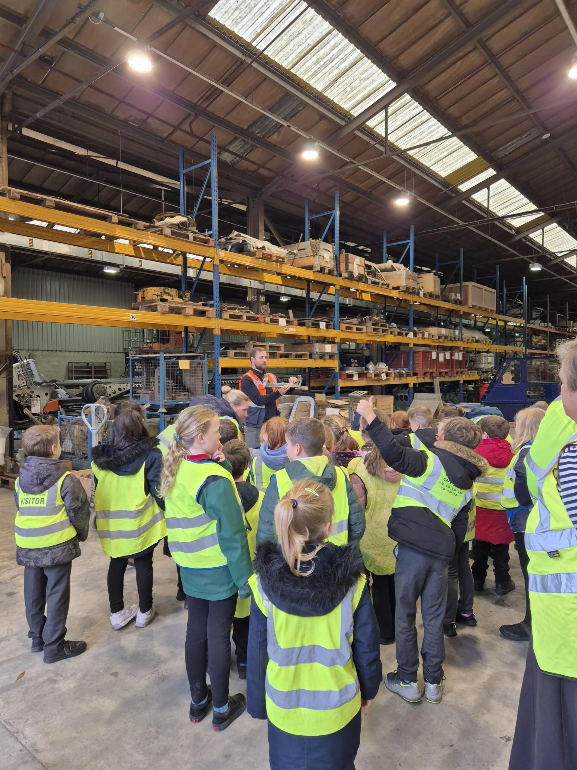 A group of children wearing yellow safety vests on a factory tour, listening to a guide in a warehouse with shelves filled with equipment and parts.