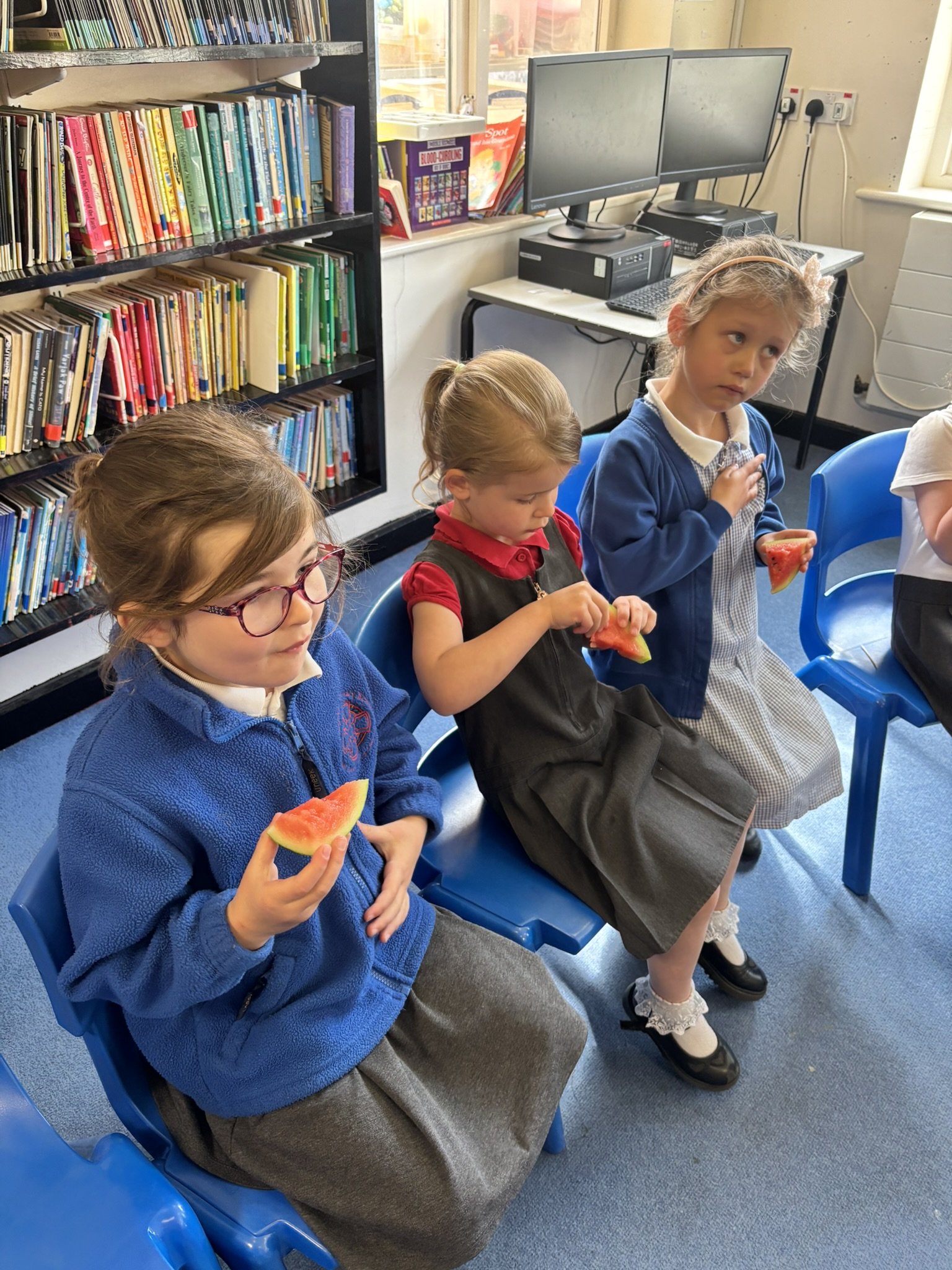 Three young girls sitting on blue chairs in a classroom, eating slices of watermelon. There are bookshelves and computer monitors in the background.