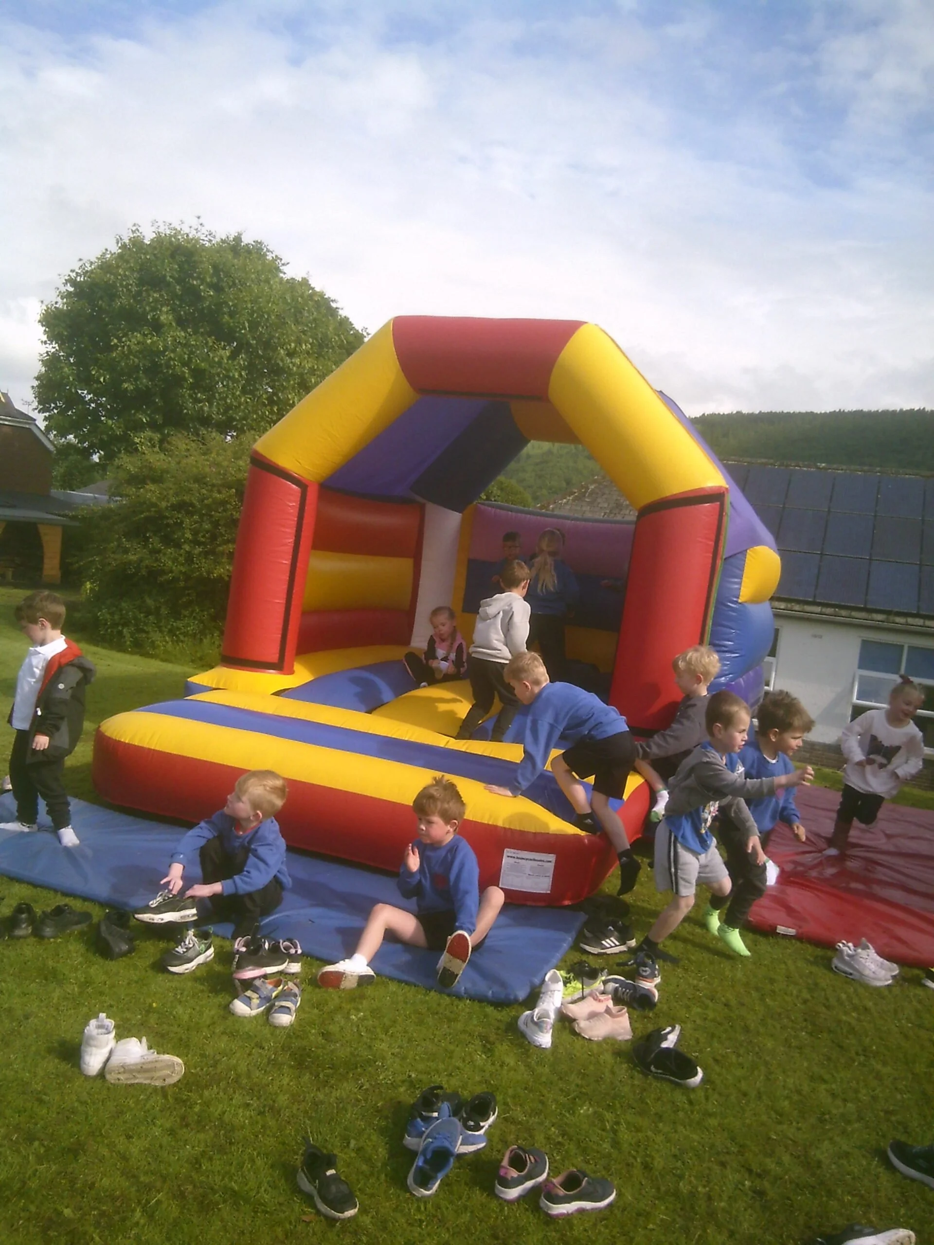Children playing on an inflatable bouncy castle outdoors, with shoes scattered on the grass around them. Blue skies with scattered clouds overhead.