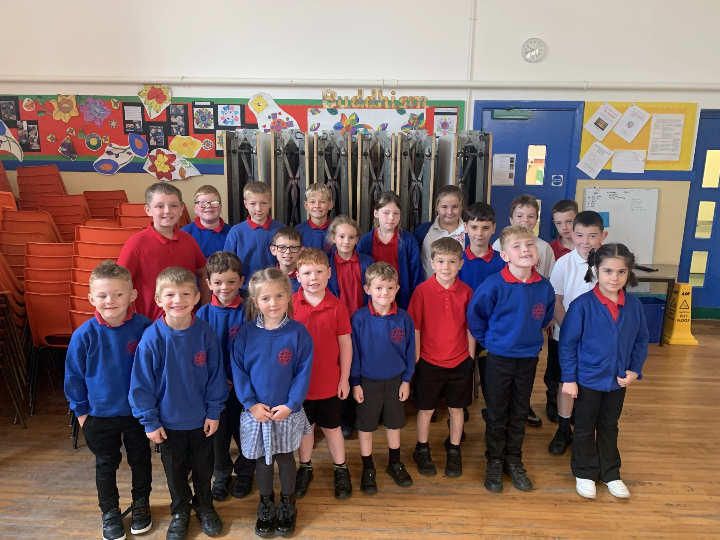 A group of school children standing together indoors in front of a decorated wall and a blue door, smiling at the camera.