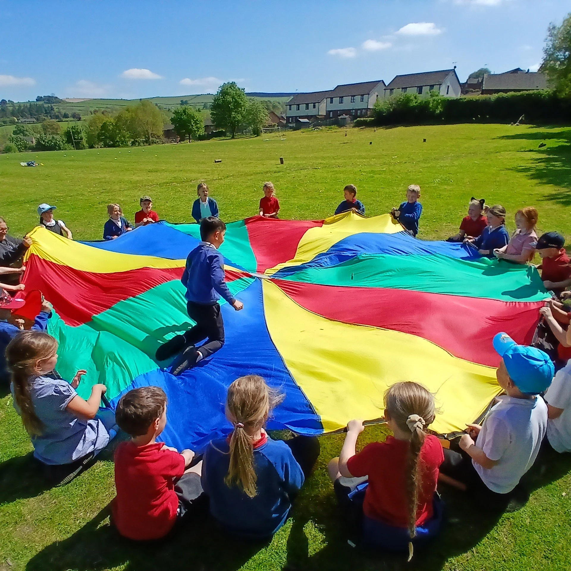 Children and adults holding a multicolored parachute outdoors on a sunny day, with a grassy field and houses in the background.