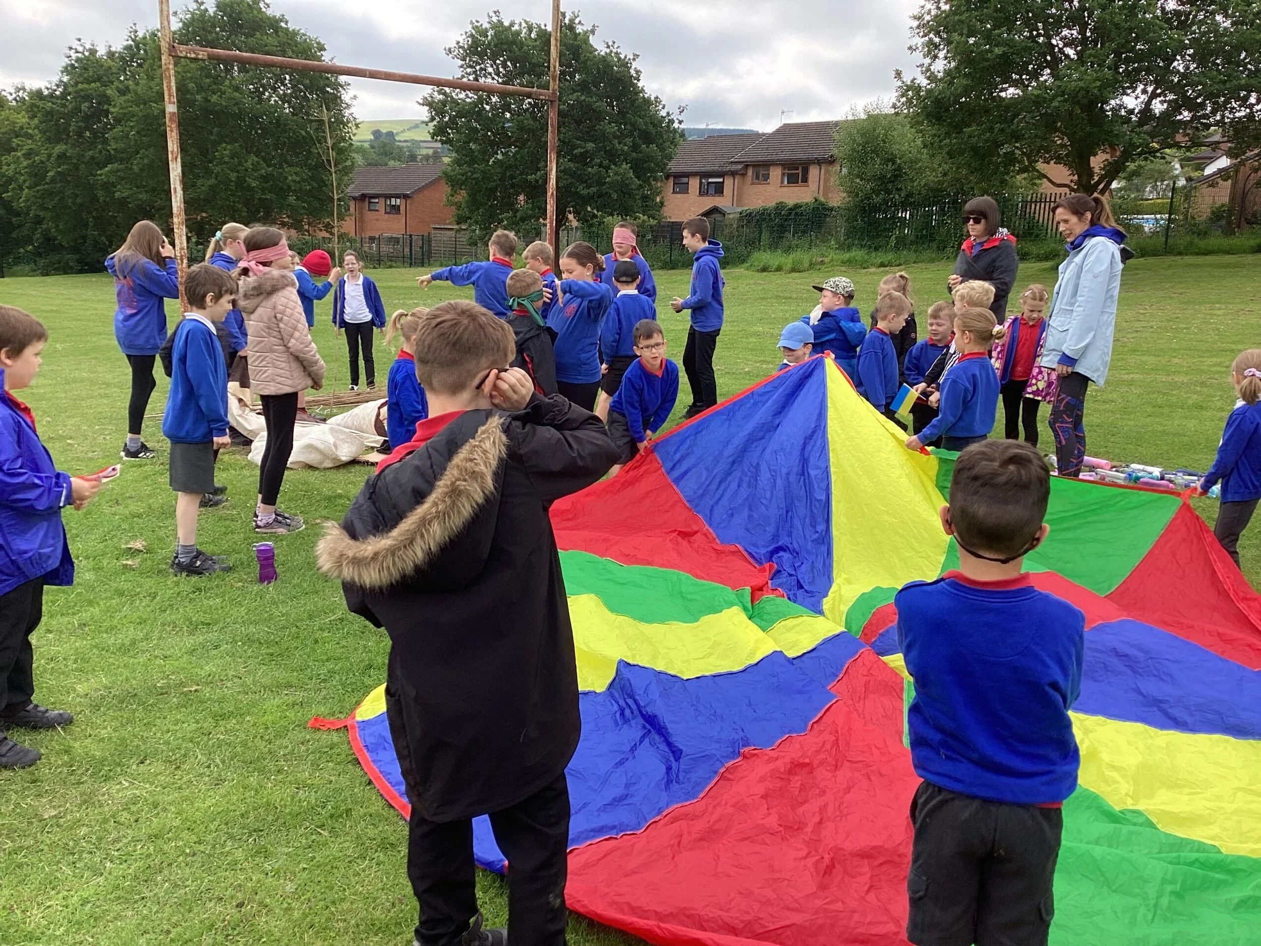 Children and adults on a grassy field, participating in a group activity involving a colorful parachute. The sky is overcast, and there are trees and houses in the background.